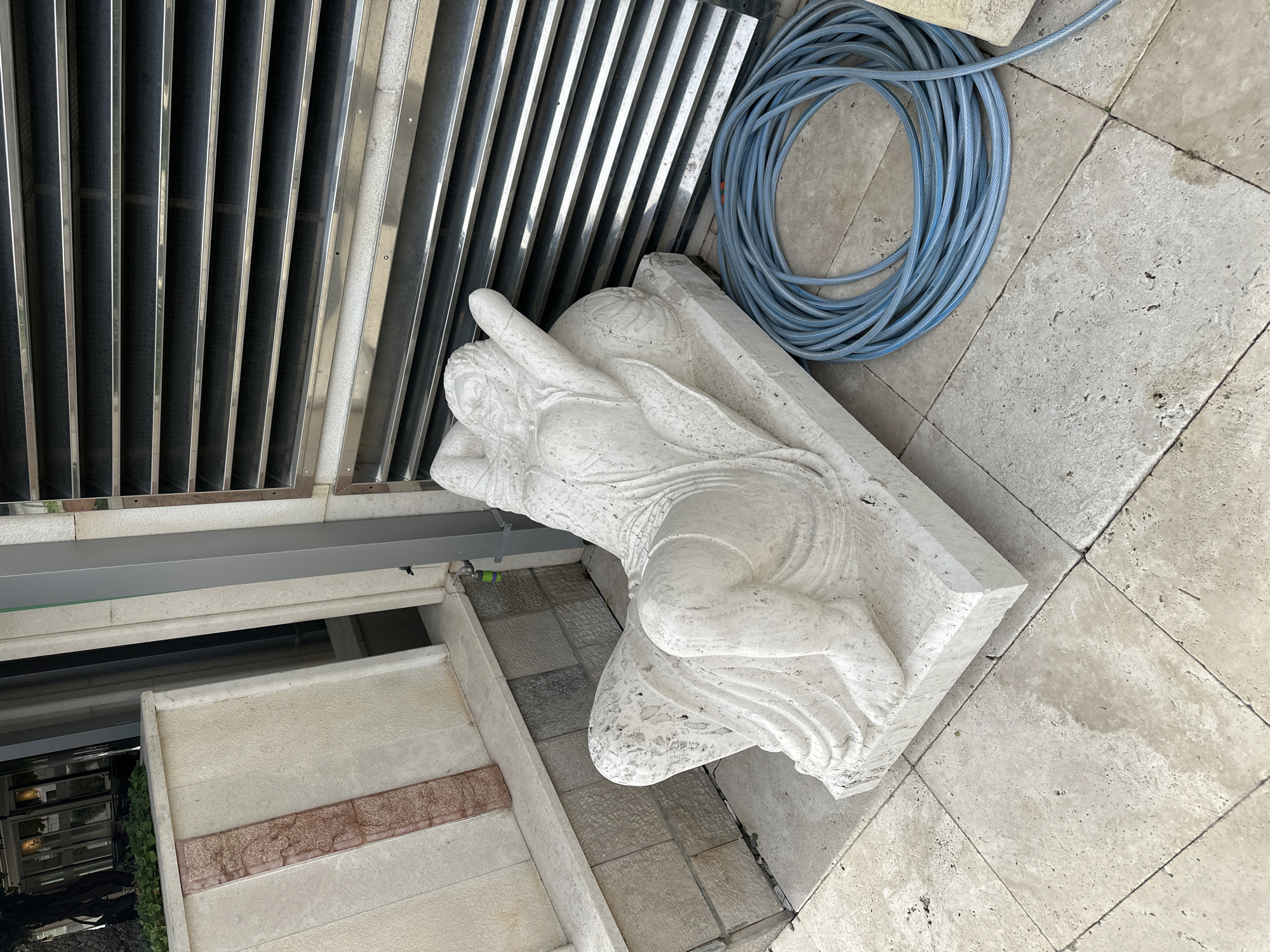 The image shows a white, sculpted marble statue of a reclining figure lying on its side on a stone-tiled surface. A coiled blue water hose is next to the statue. To the left of the statue are vertical metal grates and a wall of stone tiles with a horizontal reddish band. The location is Boreti, Montenegro.