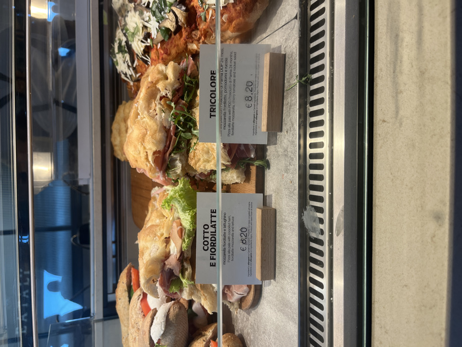 This image captures a close-up view of a refrigerated display case in a food establishment, likely a deli, cafe, or bakery, in Dorno, Italy. The case holds several freshly prepared food items, primarily focaccia or pizza alla pala sandwiches.
In the foreground and midground, two distinct sandwich types are prominently displayed, each with an accompanying white price tag and wooden stand. The top sandwich, labeled "TRICOLORE," features prosciutto di Parma, fiordilatte mozzarella, cherry tomatoes, and rocket salad. The sandwich below it, labeled "COTTO E FIORDILATTE," contains cooked ham, fiordilatte mozzarella, and lettuce. Both items are priced at € 8,20. The descriptions on the tags are in Italian and English, detailing the ingredients of each "Pizza alla pala" (paddle pizza or flatbread).
Behind these items, a large, rectangular pizza or focaccia with a reddish topping (possibly tomato sauce and other ingredients) is visible. The display case itself is made of glass and stainless steel, with reflections visible on the left side, including what appears to be part of the word "CATALI" and a blurred reflection of a person. The setting suggests a casual dining or takeaway environment where fresh Italian specialties are offered.