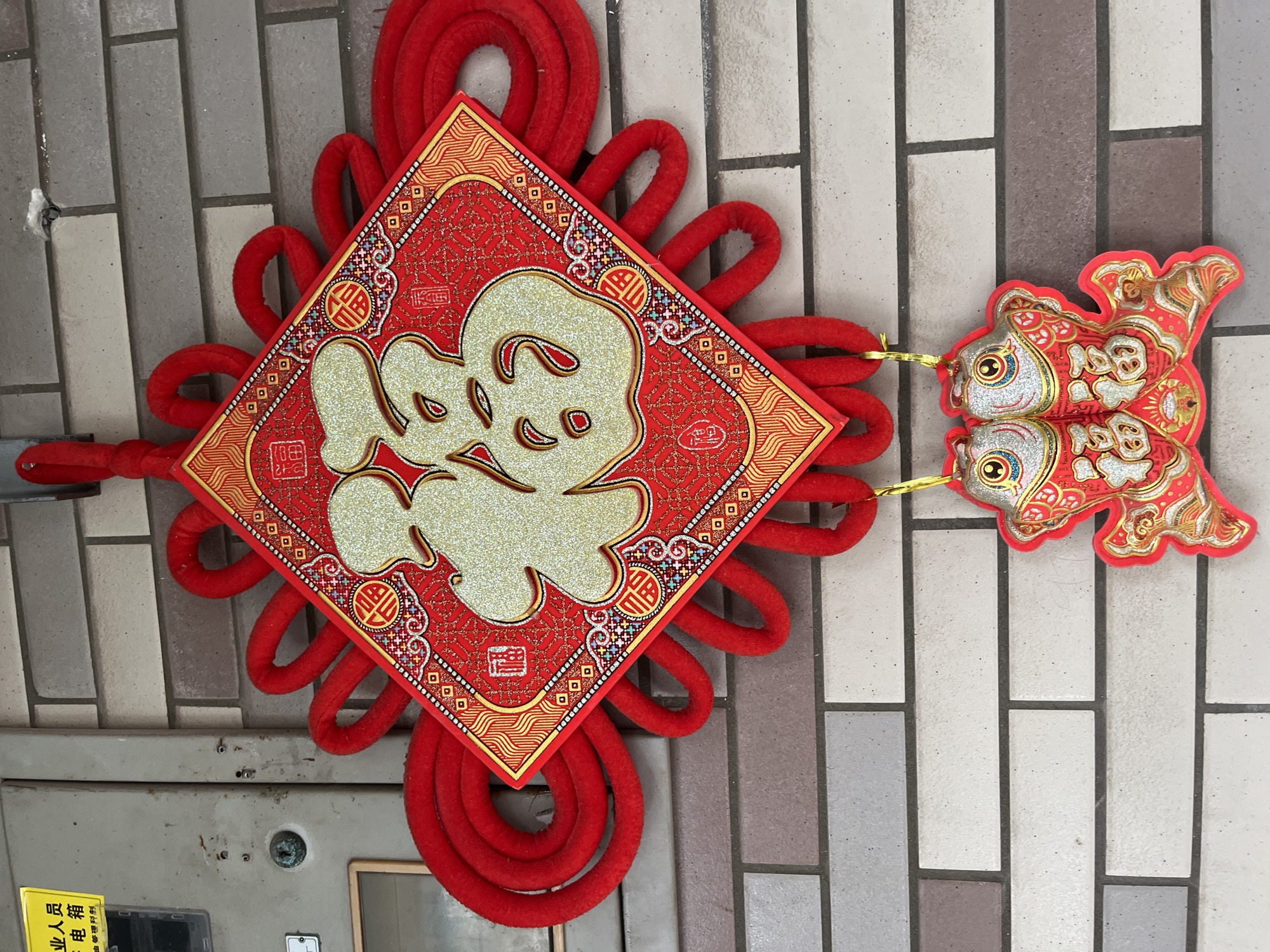 A close-up, eye-level outdoor shot captures a red Chinese knot hanging on a brick wall. The knot, adorned with a diamond-shaped red and gold panel featuring large, glittery gold Chinese characters meaning "fortune" or "blessing," is the central focus. Below the main knot, two red and gold fish-shaped ornaments with glittery details and the same characters are suspended. The brick wall, composed of light grey and beige rectangular bricks, forms the background. To the left of the knot, a light grey electrical box is partially visible, with a yellow sticker bearing Chinese text. The lighting suggests it is daytime, with no harsh shadows. No people are present in the frame. The scene is static and appears to be decorative, likely for a festival or as a cultural display.