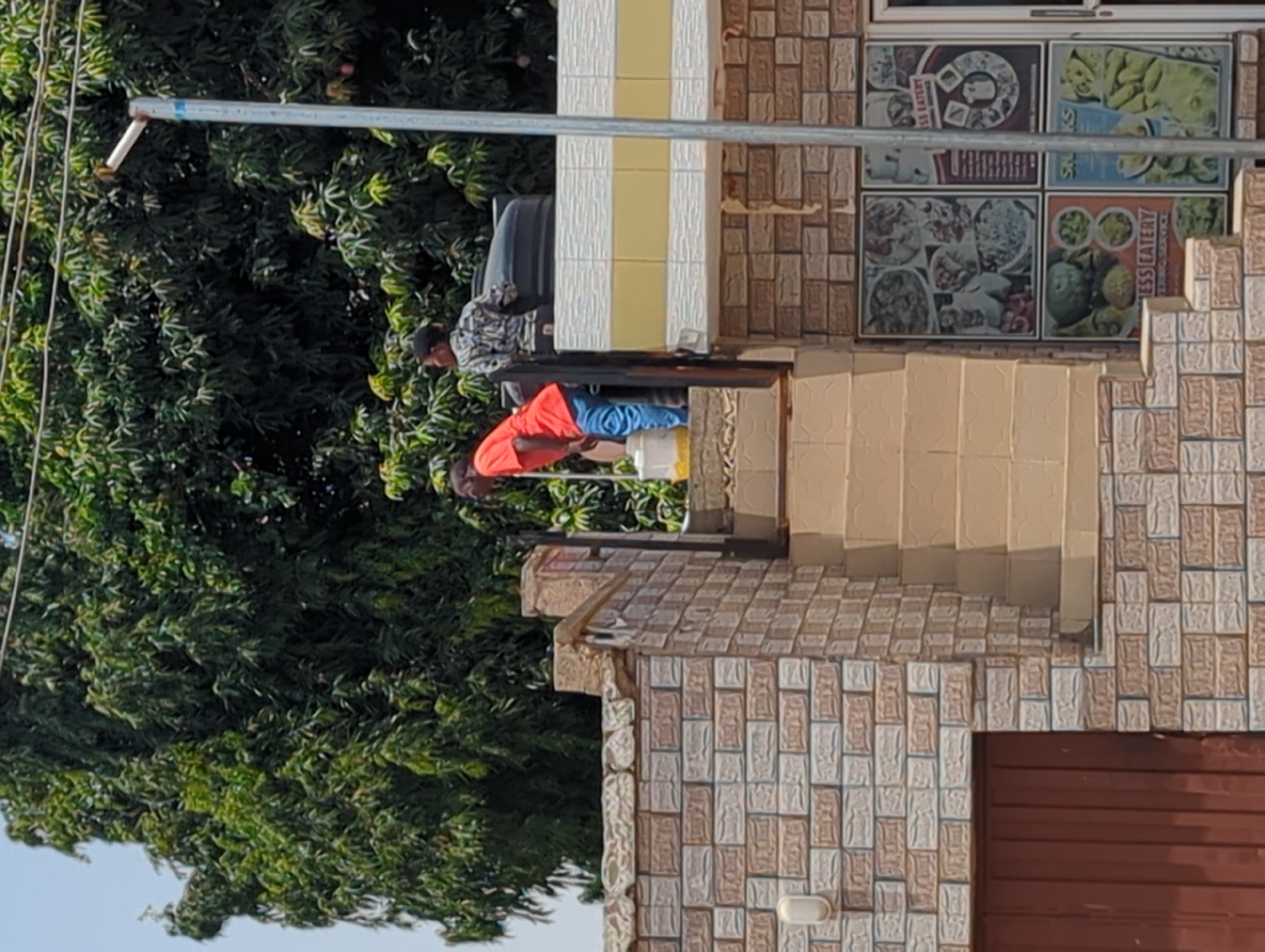 Scene in Narayi, Nigeria: Two men are present. One man, wearing an orange shirt and blue pants, is bent over near a white bucket. Another man stands behind a gray water tank, wearing a patterned shirt and dark headwear. They are situated on a raised area near the entrance of a building. The building has beige and brown tilework, visible steps, and a poster with food images displayed in the window. A large tree is adjacent to the building, with the blue sky appearing in the background. A metal pipe runs horizontally across the top portion of the image.