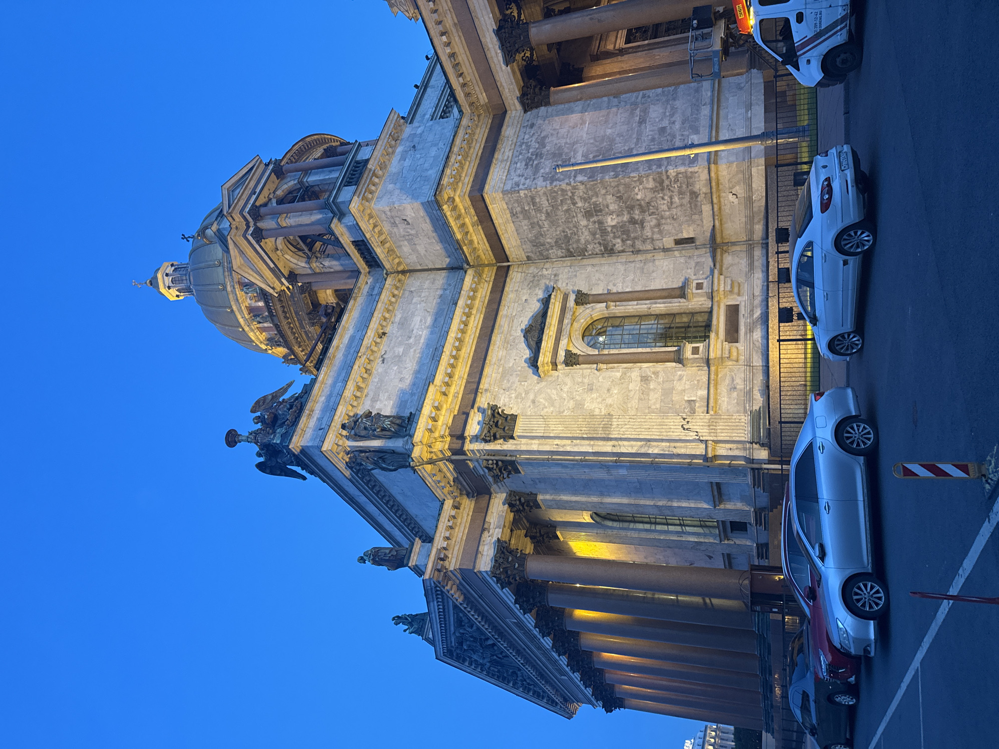 The image depicts the St. Isaac's Cathedral in Saint Petersburg, Russia, at dusk or dawn, as indicated by the deep blue sky. The grand neoclassical cathedral, constructed primarily of marble and granite, dominates the scene. Its prominent golden dome is visible at the top, topped with a cross. Sculptural elements and columns adorn the facade, illuminated by artificial lighting which casts warm yellow tones on the stone.
In the foreground, several cars are parked. From left to right, there is a dark-colored sedan, a red sedan, a silver sedan, and a white sedan. To the far right, a white van with an orange light on top is parked. The street in front of the cathedral is paved, and a metal fence runs along the side of the building. The overall scene is one of urban architecture and transportation under a twilight sky.