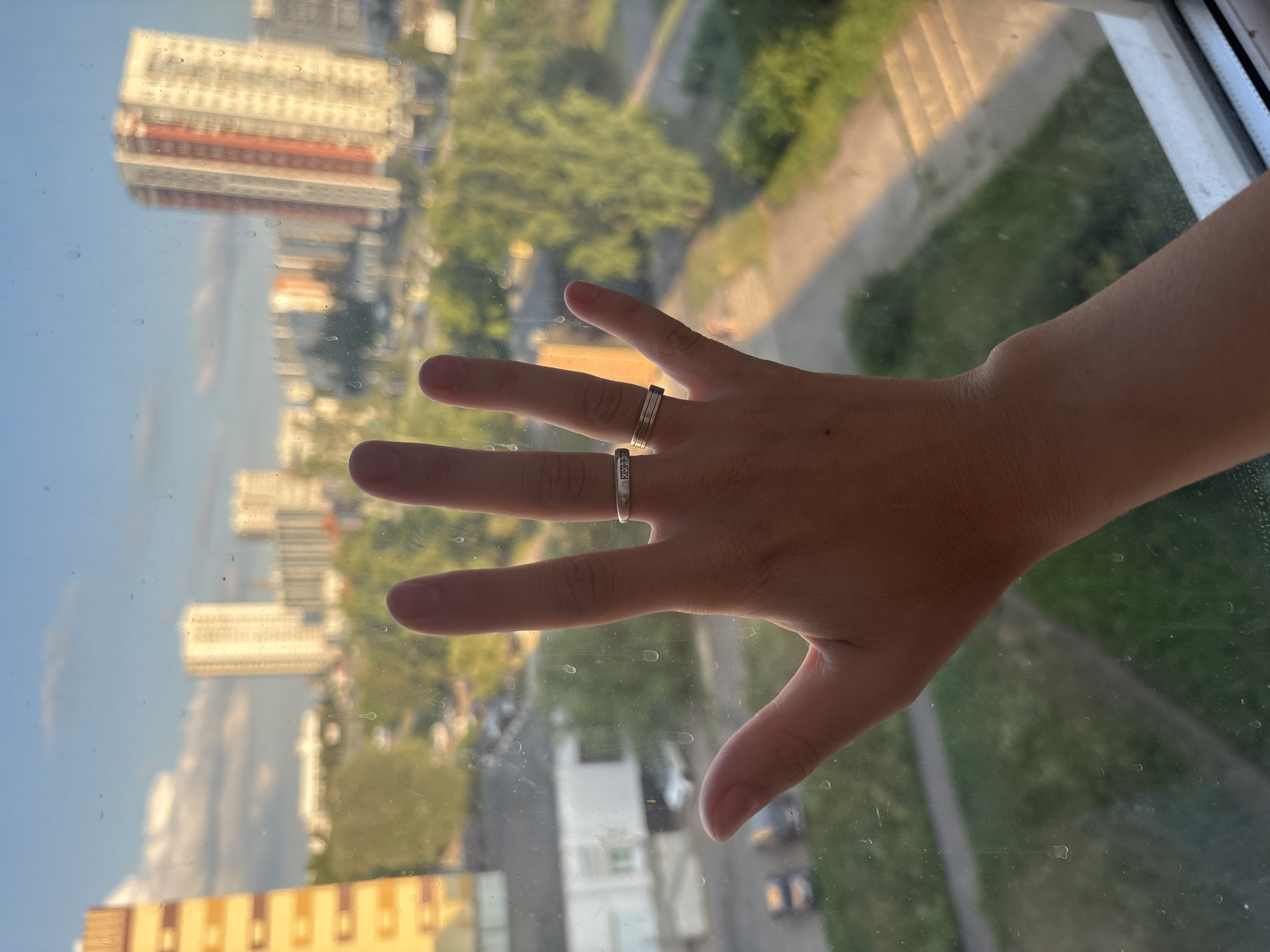 A human hand with two rings is pressed against a window. The window is dirty and shows condensation marks, suggesting it is an exterior view. Through the window, a cityscape is visible, characterized by apartment buildings and trees. The buildings appear to be a mix of residential high-rises, with some displaying architectural details like balconies and colored facades. The sky is blue with some white clouds, and the lighting suggests it is either early morning or late afternoon. The environment is urban, with hints of a street or pathway in the foreground. The crops indicate that the hand is positioned to obscure a portion of the view, and the rings are a prominent detail. It is not possible to definitively identify the location as Naberezhnye Chelny, Russia based solely on the visual information provided in the image.