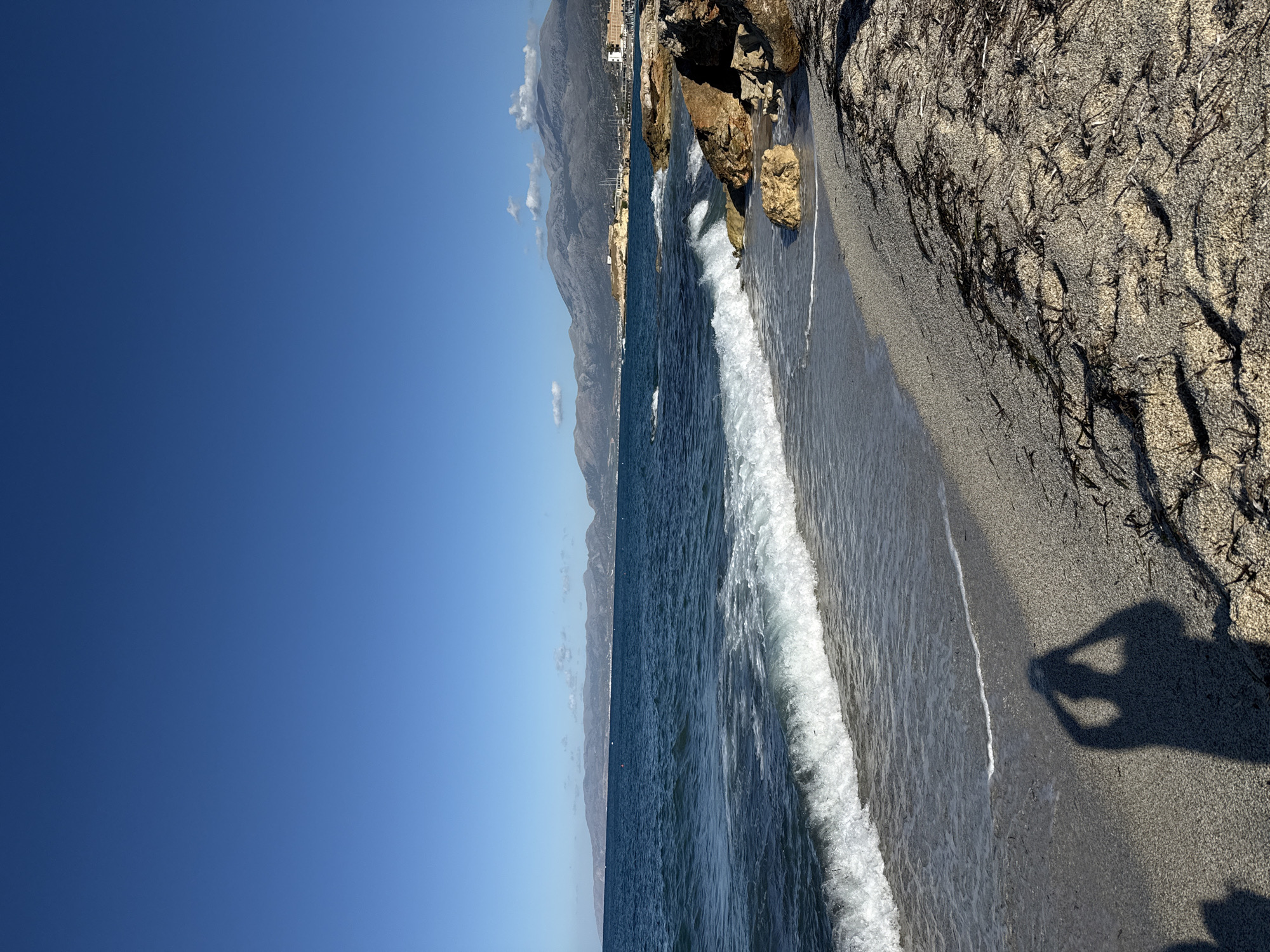 The image depicts a coastal scene in Chersonisos Municipal Unit, Greece. The foreground features a sandy beach with scattered seaweed. A shadow of a person holding a camera is visible on the sand, suggesting the photo was taken by the user. Waves are breaking on the shore, with white foam washing up onto the wet sand. In the mid-ground, the deep blue sea stretches towards a distant coastline characterized by mountains under a clear blue sky. Rocks are present along the edge of the water. The horizon shows a faint outline of land and some structures, possibly buildings or a port. The overall environment is sunny and serene.