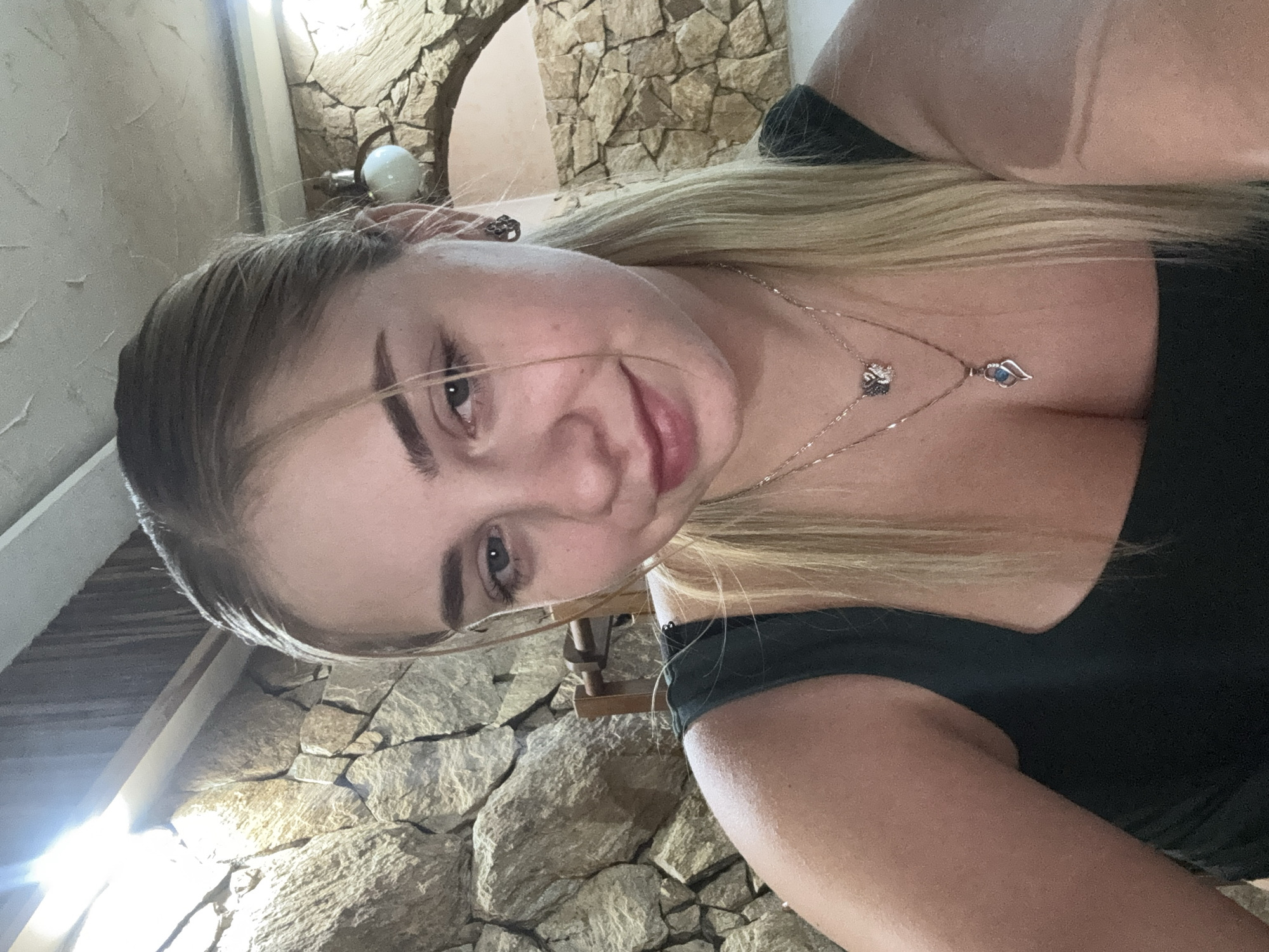 This is a medium shot selfie of a young blonde woman with her hair pulled back in a ponytail. She is wearing a dark green tank top and two necklaces. The background appears to be an interior with a stone wall. The image does not provide any specific location information for Sredets, Bulgaria.