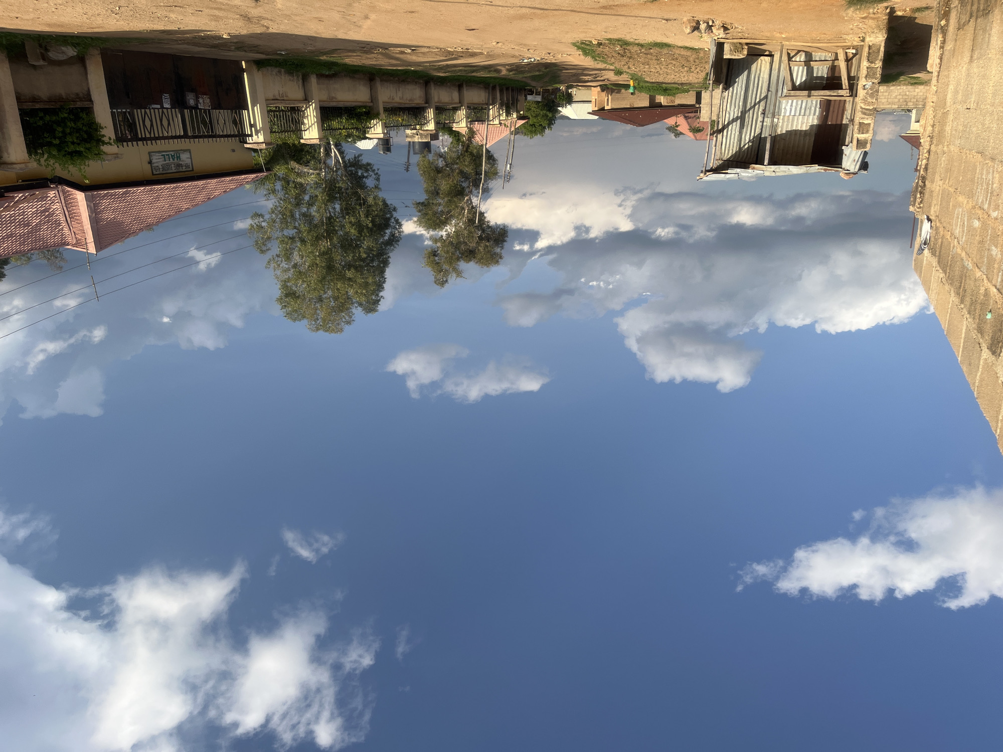 The image depicts a residential street scene in Barakin Akawo, Nigeria, under a sky with both clear blue patches and scattered cumulus clouds, some brightly lit and others appearing darker. The lighting indicates strong daylight, casting soft shadows.
The foreground features an unpaved, reddish-brown dirt road or path that extends into the distance, showing signs of wear from traffic.
On the left, a tall, unrendered concrete block wall, light brown in color, occupies the extreme left of the frame. Adjacent to it, a lean-to structure constructed from corrugated metal sheets and wooden supports stands on the dirt. This structure has an open front revealing a rudimentary wooden table or counter inside. Beyond this, other residential buildings with reddish-brown roofs are partially visible.
On the right side of the road, a series of fenced properties are observed. The most prominent property in the midground features a light yellow house with a reddish-brown tiled hip roof. Above its dark metal gate, a sign is affixed, which reads "PRS-QURANIC ACADEMIC ISLAMIC HALL". The property is enclosed by a cream-colored concrete fence topped with dark metal railings. Behind this fence, two tall trees with light-colored trunks and green foliage are prominent. Utility poles and overhead wires run along this side of the road, and a black water storage tank on a stand is visible behind a fence.
In the far right background, a small human figure, possibly a child, wearing a yellow top, is discernible near a low embankment. The overall scene conveys a semi-urban or developing residential environment.