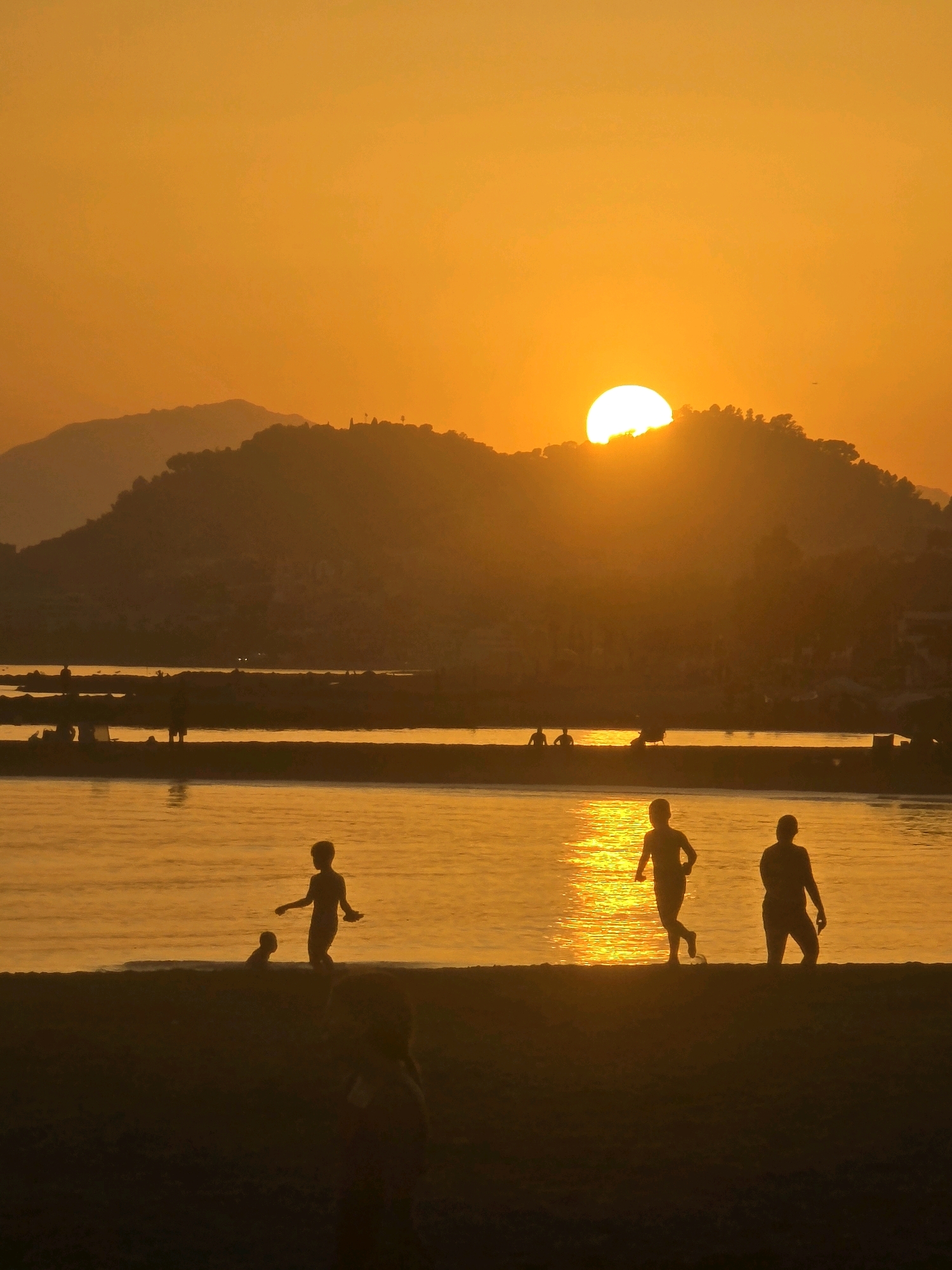 A vertical, low-angle, silhouette shot captures a vibrant orange sunset over a body of water, likely the Mediterranean Sea near Málaga, Spain. The sun, a bright, glowing orb, is positioned on the right side of the frame, partially obscured by a distant, tree-covered mountain range. The water's surface reflects the sunset's golden hues, creating a shimmering effect.
In the foreground, a dark silhouette of a beach is visible, with three children playing near the water's edge. One child, to the left, is partially submerged in the water. Another child, in the center, is running towards the right, with one leg extended as if mid-stride. To the right, a third child stands still, facing left. Further back, silhouetted figures of other people can be seen on a pier or breakwater that stretches across the middle ground of the image. The overall atmosphere is one of serene, late-day recreation.