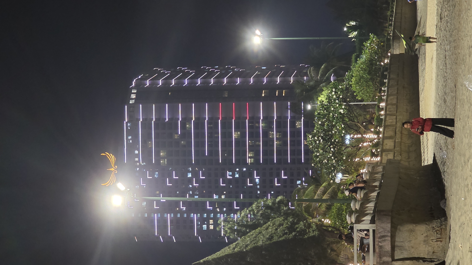 The image displays a nighttime scene on Nha Trang Beach, Vietnam. In the background, a tall, multi-story high-rise building with a distinctive golden winged emblem at its apex is prominently featured. The building's facades are illuminated by numerous horizontal purple LED light strips, with several red strips visible on its right side. A bright streetlamp stands in the foreground of the building, partially obscuring the golden emblem.
A concrete promenade with decorative balusters and lush tropical vegetation, including palm trees and flowering frangipani trees, spans the mid-ground, separating the high-rise from the sandy beach. Several individuals are present on this promenade: one person is seated on a bench, observing a device, while others stand near the balustrade.
In the foreground, on the sandy beach, a woman wearing a red long-sleeved top and dark pants stands, looking down at a mobile phone held in her hands. To her right, a young child in a green shirt and shorts moves across the sand, possibly holding a light green object, which could be a kite. The scene is illuminated by streetlights and the building's architectural lighting under a dark night sky.