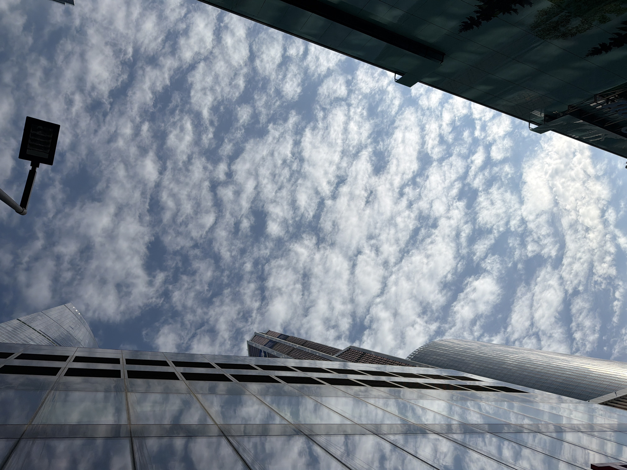 The image is a low-angle view towards a cloudy sky, framed by building structures. Several modern skyscrapers rise into the frame from the bottom, featuring glass facades that reflect the sky. A dark, rectangular street light is positioned on the left side. Above, the edges of other buildings cut into the top of the frame. The location is within the Moscow International Business Center (MIBC), also known as Moscow-City, in Moscow, Russia.