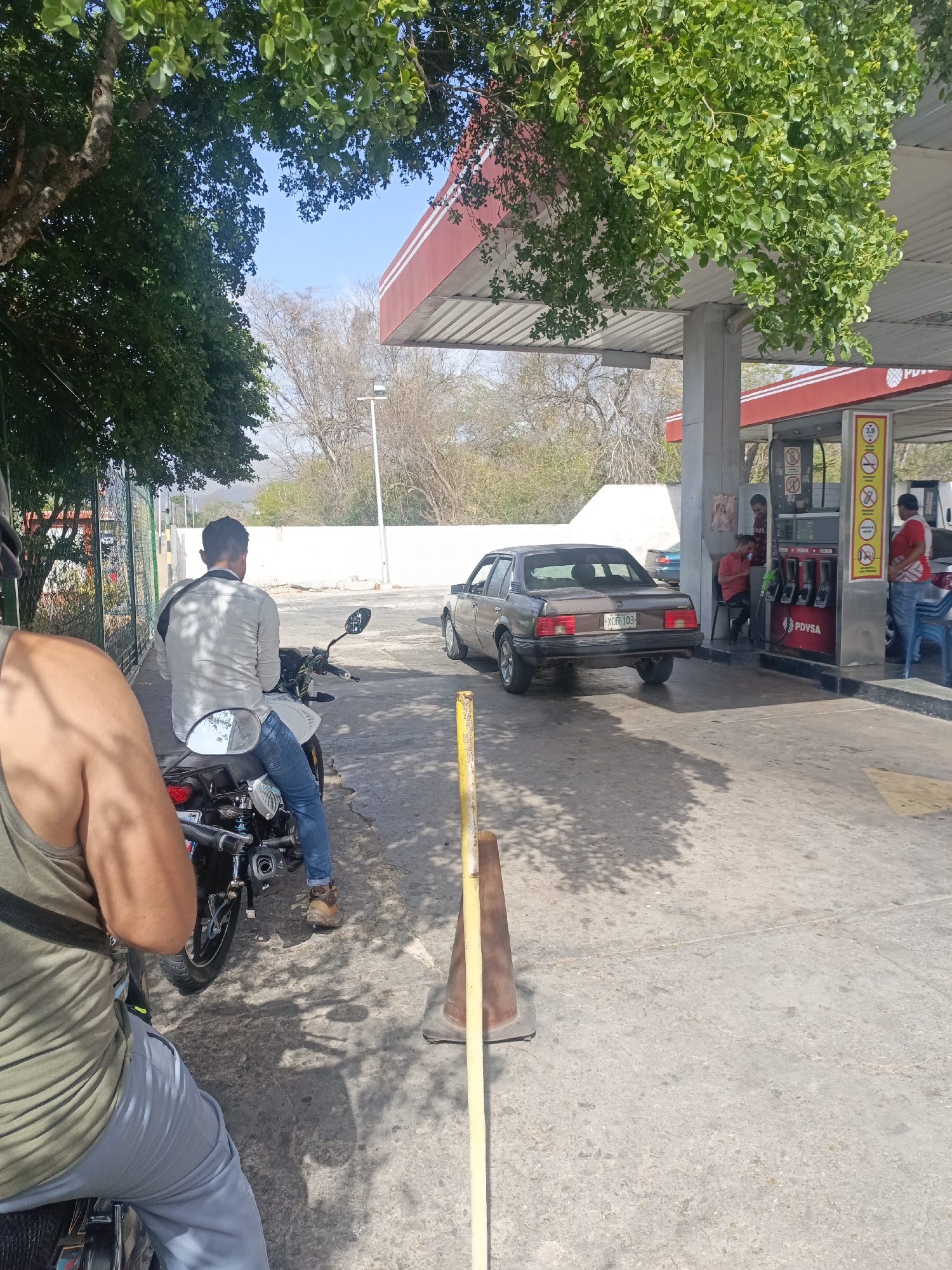 The image depicts a gas station in what appears to be La Tagua, Venezuela, evident from the PDVSA signage. A dark-colored sedan, displaying license plate "XDR-103," is parked at one of the pumps. A man in a red shirt is standing next to the gas pump, while two others can be seen behind the pumps. Two men are on motorcycles, waiting in line. A yellow pole and cone are near the waiting line. The surrounding environment consists of trees and a white wall. The weather appears sunny.