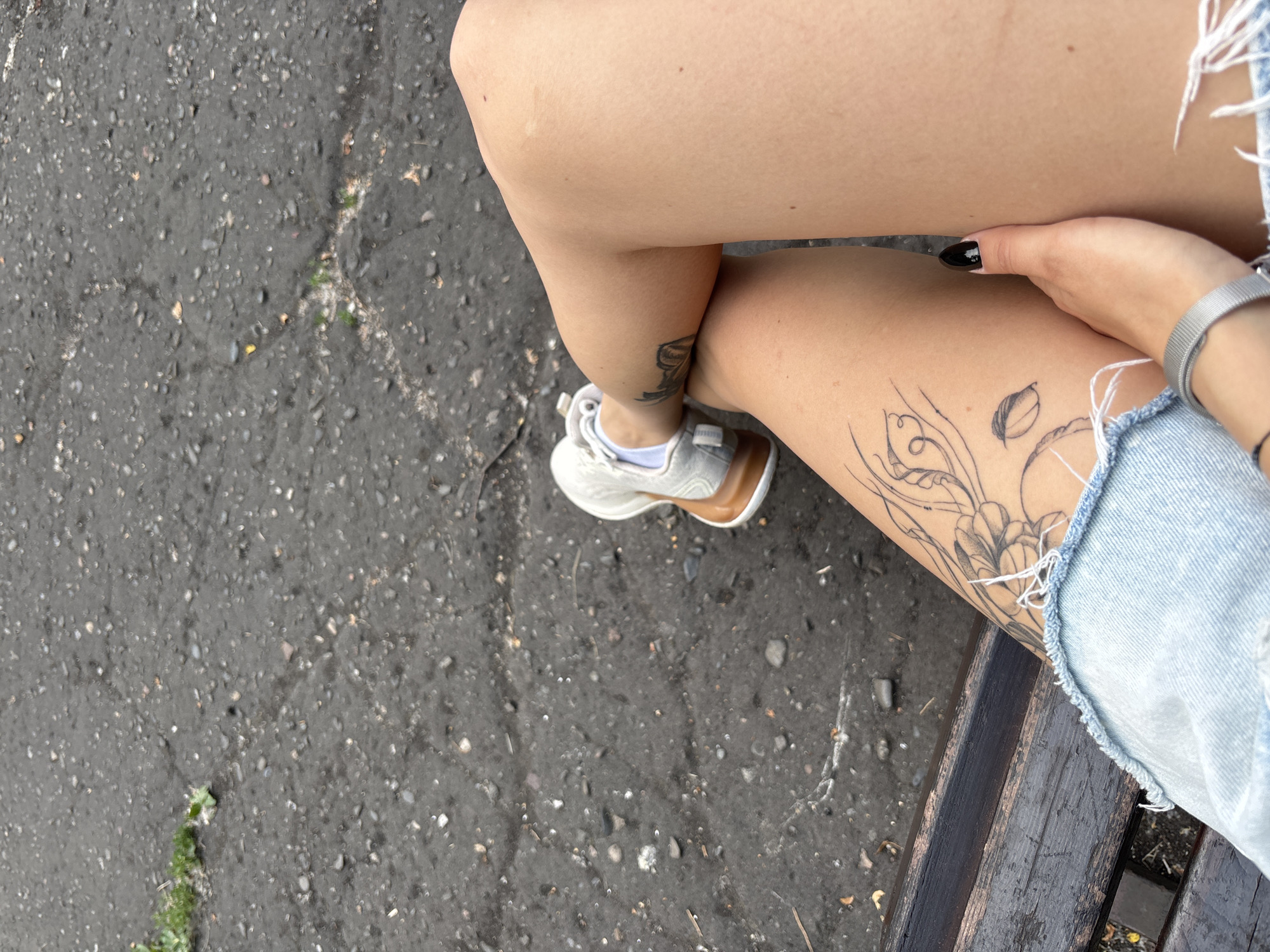 The media file shows a close-up of a person's legs and feet, resting on a dark wooden bench. The ground is asphalt with small stones and some patches of green grass. The person is wearing a pair of white sneakers with brown accents and light blue denim shorts. There are two tattoos visible on the person's left leg: one is a stylized image of a flower, and another, partially visible, appears to be a depiction of a winged creature or insect. The person's right hand, with black nail polish, is resting on their leg. A silver watch is visible on their wrist. The image is taken from a high angle, looking down at the legs. The lighting suggests it is daytime, likely outdoors. There is no visible text in the image. The setting appears to be an outdoor urban area, possibly a park or a public space, given the bench and the asphalt ground. The overall mood is casual and relaxed, suggesting a moment of rest. Given the location context of Novokuznetsk, Russia, the scene depicts a person enjoying a moment outdoors in what appears to be a temperate climate.