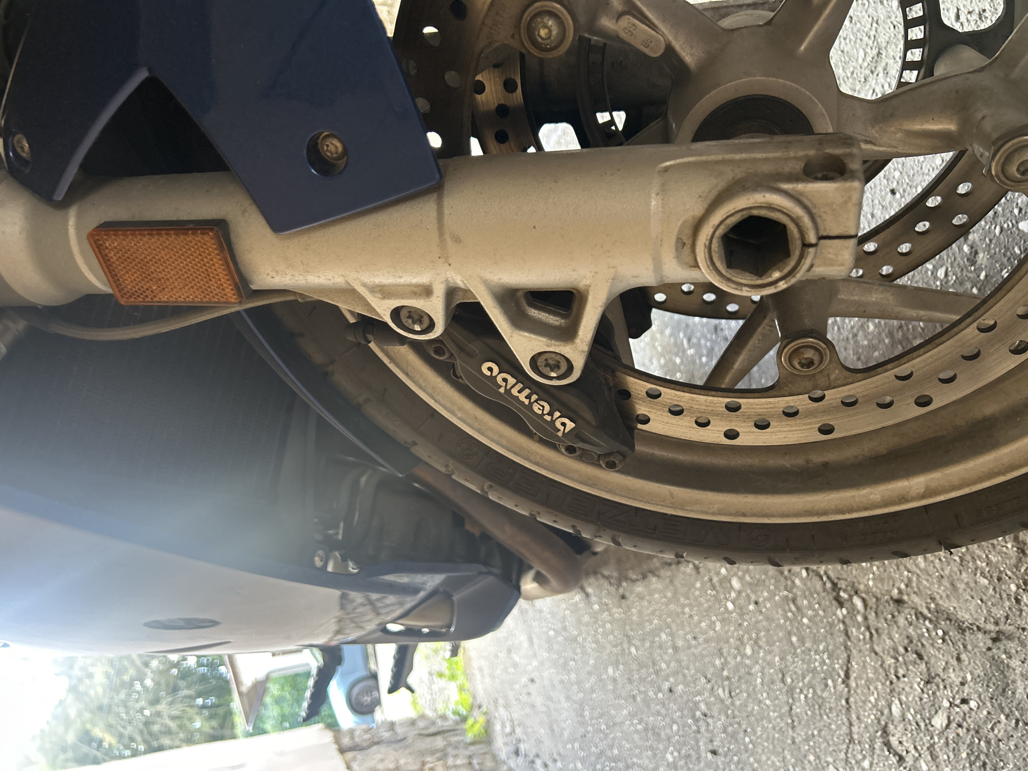 The image shows a close-up, low-angle view of the front wheel and suspension of a motorcycle. The motorcycle is blue and appears to be parked on an asphalt surface. The focus is on the front brake caliper, which is branded "Brembo", and the perforated brake disc. The motorcycle's front fork is visible, along with a portion of the tire and wheel rim. In the background, there is a stone wall and some greenery, suggesting an outdoor setting. A light blue car is partially visible in the distance. The overall lighting suggests it is daytime. There are no people or specific actions depicted. The scene is a static representation of a parked motorcycle. The image does not contain any visual elements that indicate the location is specifically in Vilnius, Lithuania.