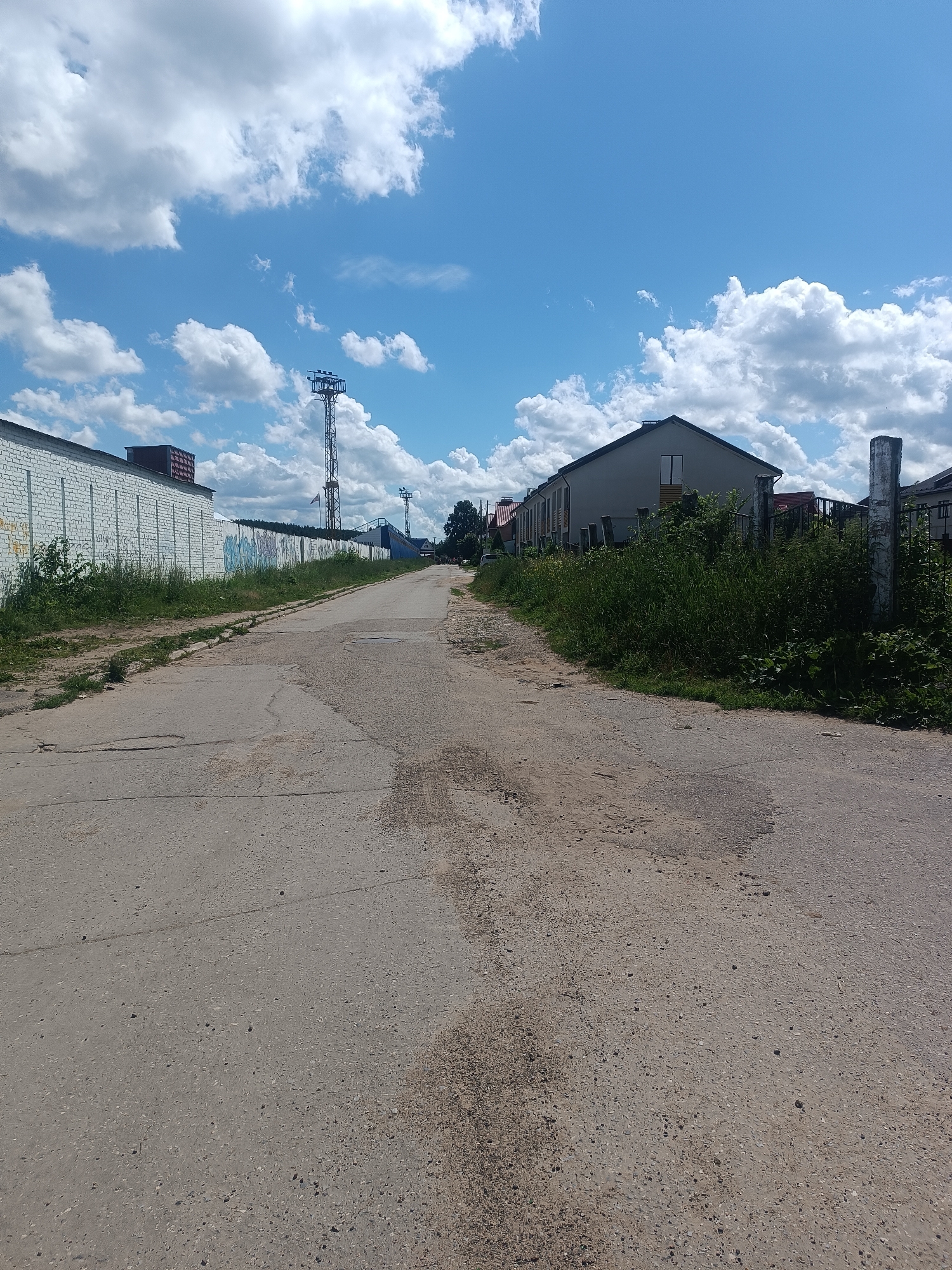 The image depicts a paved road in a semi-urban or industrial area, likely in Chaikovsky, Russia, given the context. The road surface is worn and shows patches of repair and discoloration. On the left, a long, white brick wall with graffiti runs alongside the road. Beyond the wall, a red-roofed building and a tall metal communication tower are visible. The right side of the road is lined with dense green vegetation and a few residential buildings with dark roofs and light-colored facades. A tall, weathered concrete pillar stands by the roadside on the right. The sky is bright blue with scattered white cumulus clouds, suggesting fair weather. The overall impression is of an utilitarian, perhaps developing, area.