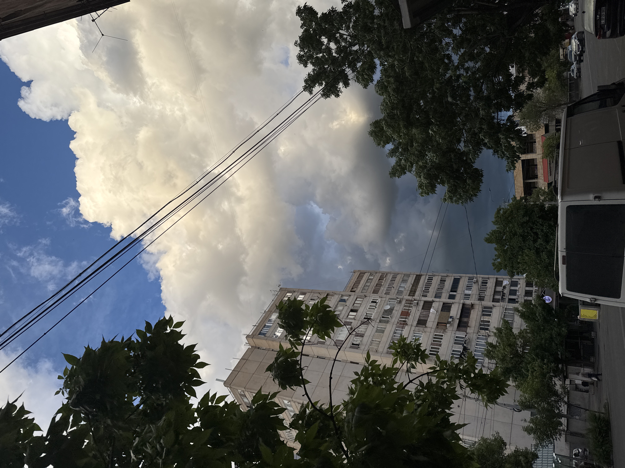 The image shows a street scene in Yerevan, Armenia. The view is upward, capturing a tall, Soviet-era apartment building with multiple windows and some satellite dishes. Trees frame the bottom of the image. A white van is parked on the right side of the frame, partially obscuring the view. Utility lines stretch across the sky. The sky is filled with dramatic clouds, ranging from white and puffy to dark and ominous. In the distance, there is an unfinished building. Cars are parked on the street. The overall impression is that of a cloudy day in an urban setting, with a mix of older and newer structures.