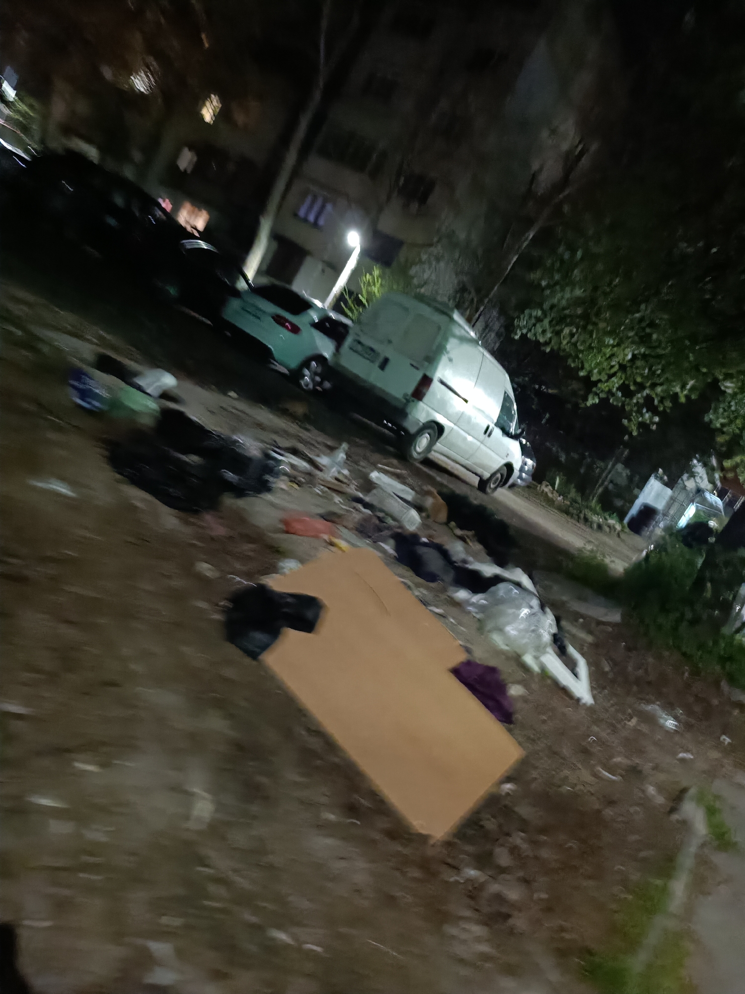 The image depicts a trash-strewn area in Pleven, Bulgaria, at night. The scene includes assorted garbage, such as plastic bags, discarded items, and a large piece of cardboard lying on the ground. In the background, cars and a white van are parked alongside a multi-story building with visible windows. Trees are present on the right side of the image. The general impression is one of neglect and littering.