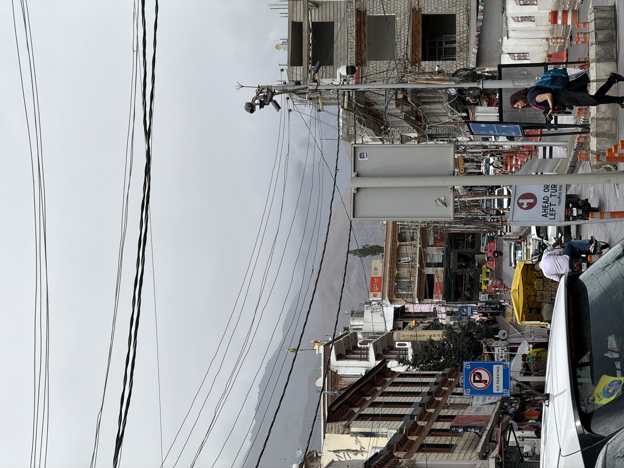 The image shows a street scene in Leh, India. The street is lined with multi-story buildings, many of which have traditional Ladakhi architecture with carved wooden details. Several power lines crisscross overhead against a cloudy sky, with hazy mountains visible in the background.
In the foreground, a white car is partially visible on the left, and a white SUV is seen behind it. A man in a white shirt is riding a motorcycle in the middle of the street. Further up the street, a woman with a blue backpack is walking on the sidewalk. There are several traffic signs, including a "No Parking" sign and a sign indicating "Ahead or Left Turn, Traffic Pole". Orange traffic cones line the road. A yellow tent is set up on the right side of the street, with boxes stacked beneath it. Various shops and businesses are visible along the street, with some displaying signs like "Hotel Disket Cha" and "Montagne". A few people are walking on the sidewalks and in the street. Security cameras are mounted on a utility pole. Prayer flags are strung across some buildings.