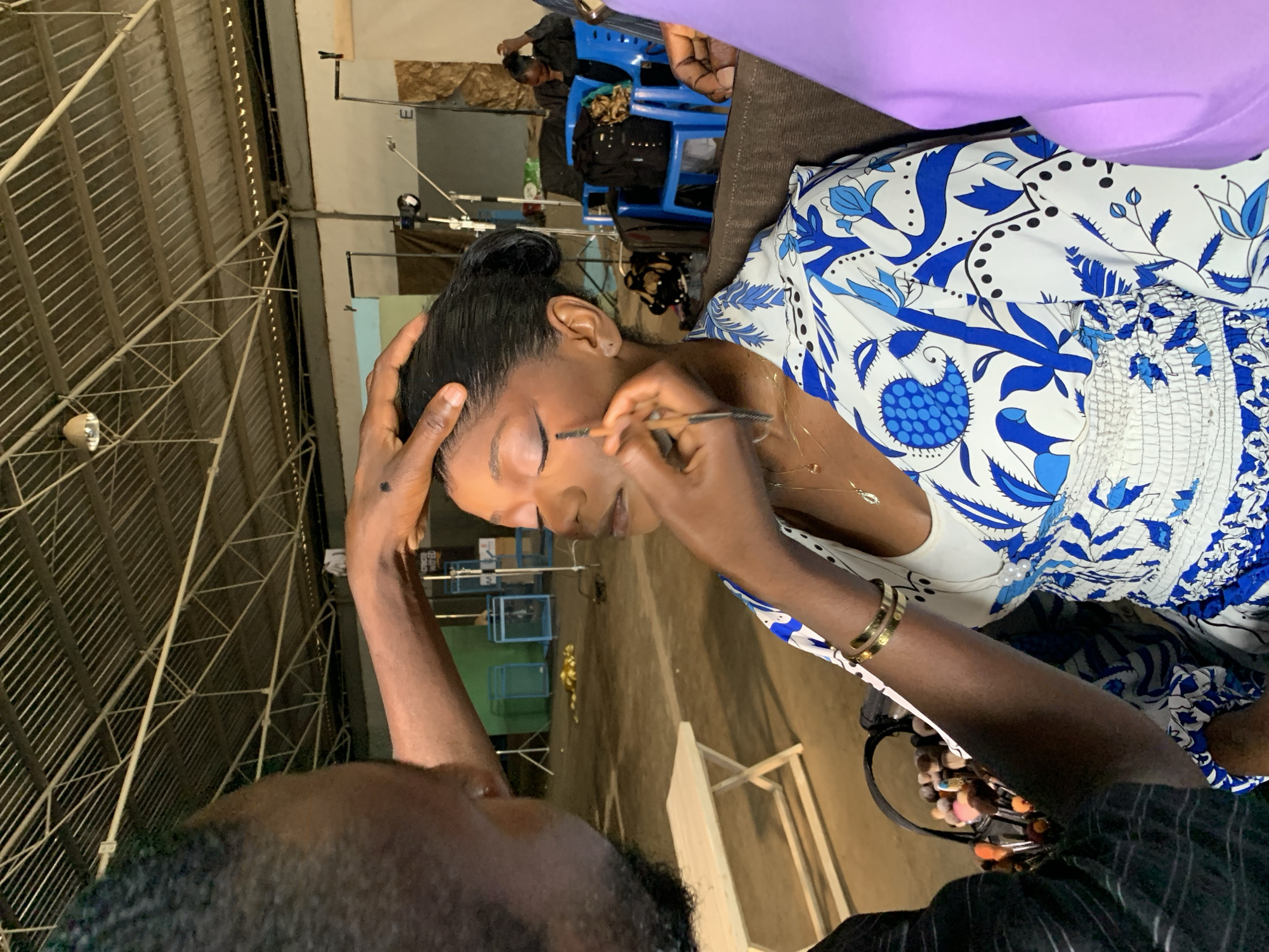 A medium shot captures a woman receiving makeup application in what appears to be an indoor, possibly industrial setting. The woman, with dark hair pulled back, is seated and looking down, her eyes closed as a makeup artist, with hands visible, applies product to her eyebrows with a brush. She is wearing a white top with a blue floral pattern and a gold bracelet on her left wrist. A portion of another person's head and shoulder, with dark hair and beard, is visible in the foreground, suggesting the photographer is also present.
In the background, a blue plastic chair is occupied by another person wearing dark clothing. Further back, a metal structure with horizontal bars, possibly part of a roof or shelving, is visible. The lighting appears to be artificial. The overall ambiance suggests a behind-the-scenes or preparation moment. The image orientation is vertical with the top of the frame on the left. There is no visible text. The location context provided is Barakin Lerawi, Nigeria.