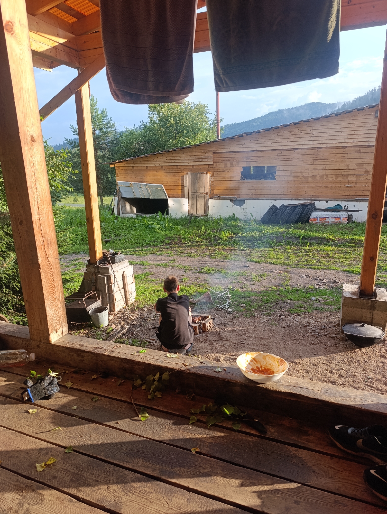 The image depicts a rustic outdoor scene, likely at a dacha or rural residence in Biryuchul, Russia. In the foreground, a wooden porch is visible, with scattered leaves and a pair of black athletic shoes. A white bowl containing remnants of food sits on a wooden beam. In the middle ground, a person is crouched down near a small barbecue, tending to a fire. Various items are scattered around the barbecue area, including a bucket and grill grates. A rustic wooden building with a large opening is situated in the background. The area surrounding the building is grassy and dirt-covered. The scene is bathed in the warm light of what appears to be late afternoon or early evening sun. The overall impression is one of a casual outdoor gathering or meal preparation in a natural, somewhat unkempt setting.