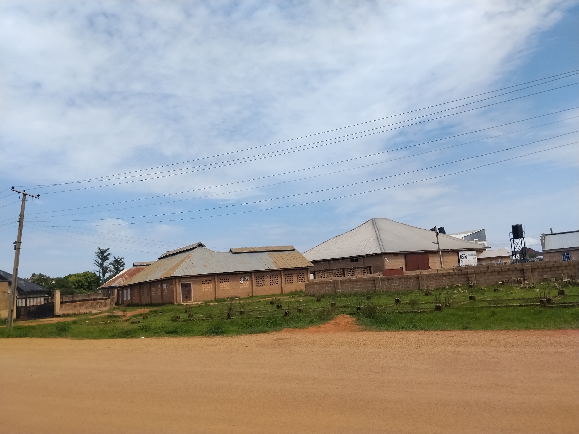 The image shows a view of buildings and infrastructure in Nabatum, Nigeria. In the foreground, there is a dirt road. Behind the road is a grassy area with some small, incomplete concrete structures. Further back are several buildings constructed from light-colored bricks with corrugated metal roofs. One building on the left has a gate and appears to be a residential or commercial property. Another larger building in the center has a distinctive A-frame roof and a series of window openings along its side. To the right, another building with a similar roof style is visible, along with a water tower on a metal frame. Overhead, power lines stretch across the frame, supported by a utility pole on the left side of the image. The sky is partly cloudy with a light blue hue. The overall scene depicts a developing area with ongoing construction and existing structures.
