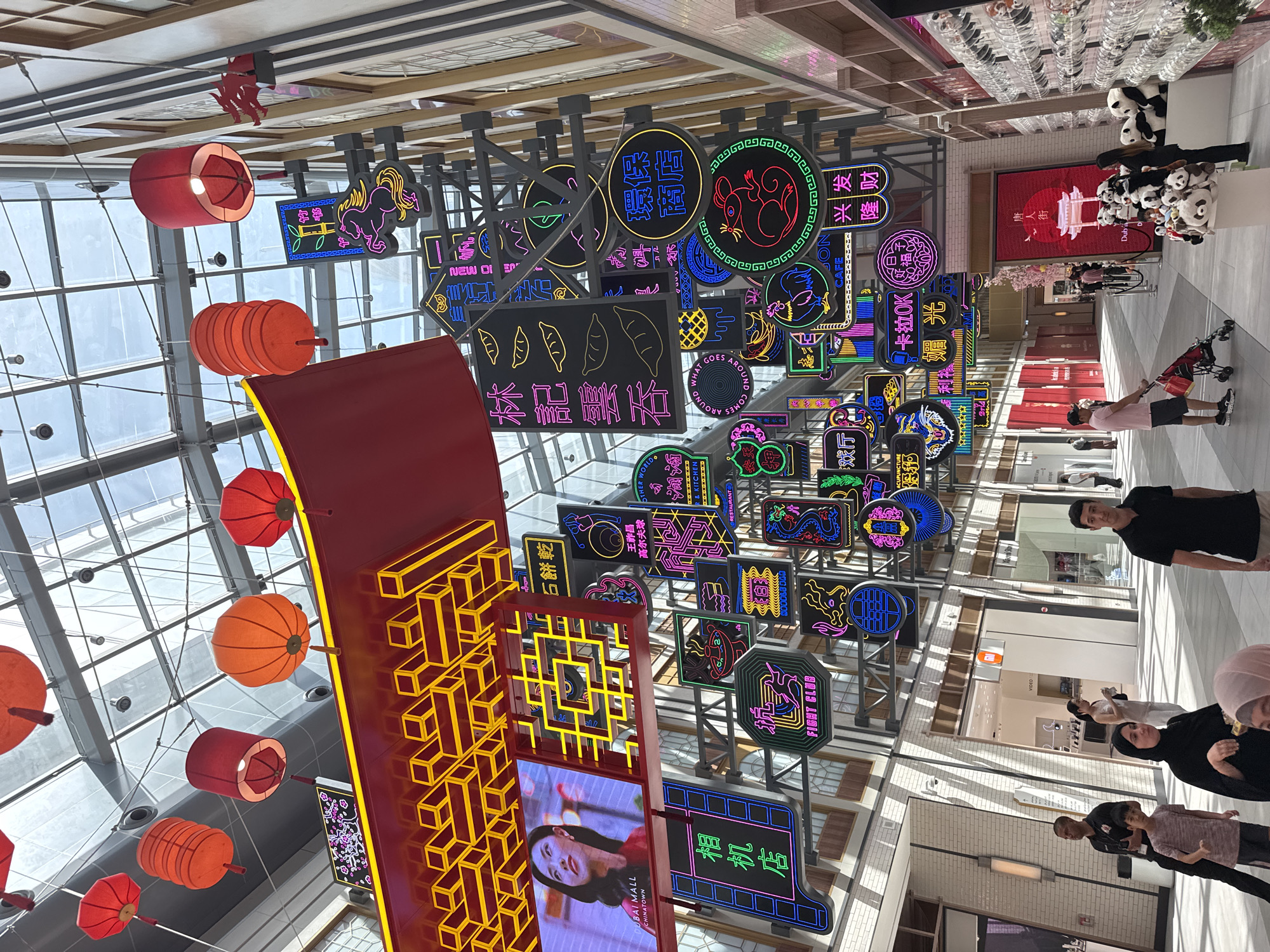 The image captures an indoor scene in the Chinatown section of the Dubai Mall, Dubai, United Arab Emirates. A complex array of illuminated signs in various shapes and colors, many featuring Chinese characters, hangs from the ceiling. The signs advertise businesses like restaurants, cafes, and services. Red and orange Chinese lanterns are suspended nearby. A large vertical display panel with a golden geometric design and an advertisement with a woman's face is also visible. Several people are in the scene, including a man in a black shirt facing the camera, and a family walking with a stroller. Merchandise, including panda plush toys, is visible in the background. The mall architecture includes a glass ceiling with a grid pattern.