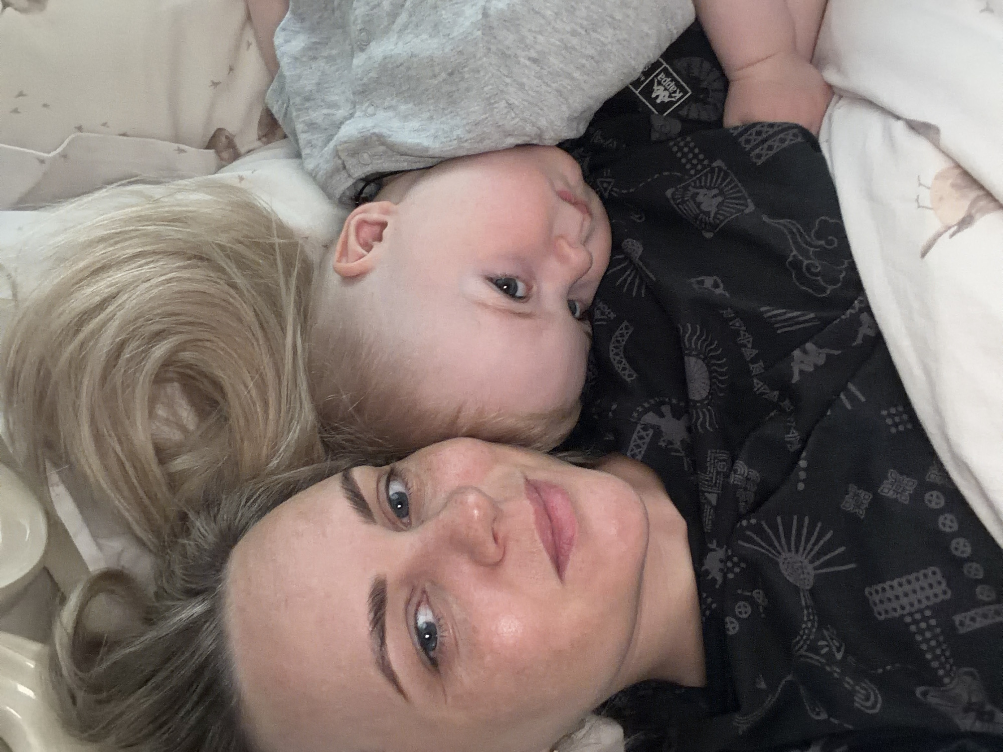 An adult woman and an infant are lying side-by-side, likely on a bed, captured from a slightly overhead perspective. The woman, with blonde hair spread out on the bedding and blue eyes, looks calmly directly into the camera. Her face shows a serene expression. Beside her, an infant with light blonde hair and blue eyes rests their head, looking slightly towards the right of the frame with a neutral, observant expression.
The adult is wearing a dark top with intricate grey patterns, while the infant wears a simple grey sleepsuit. Surrounding them are beddings: a light-colored blanket with subtle, lighter patterns (possibly birds) on the left, and a dark patterned blanket or sheet on the right, featuring various grey designs including sunbursts, clouds, and geometric shapes. A small black tag with the white "Kappa" logo is visible on the dark patterned fabric near the infant's head. The indoor setting and the subjects' relaxed postures suggest a peaceful, intimate moment, possibly in a bedroom. There are no specific visual cues in the image indicating the location context of Votkinsk, Russia.