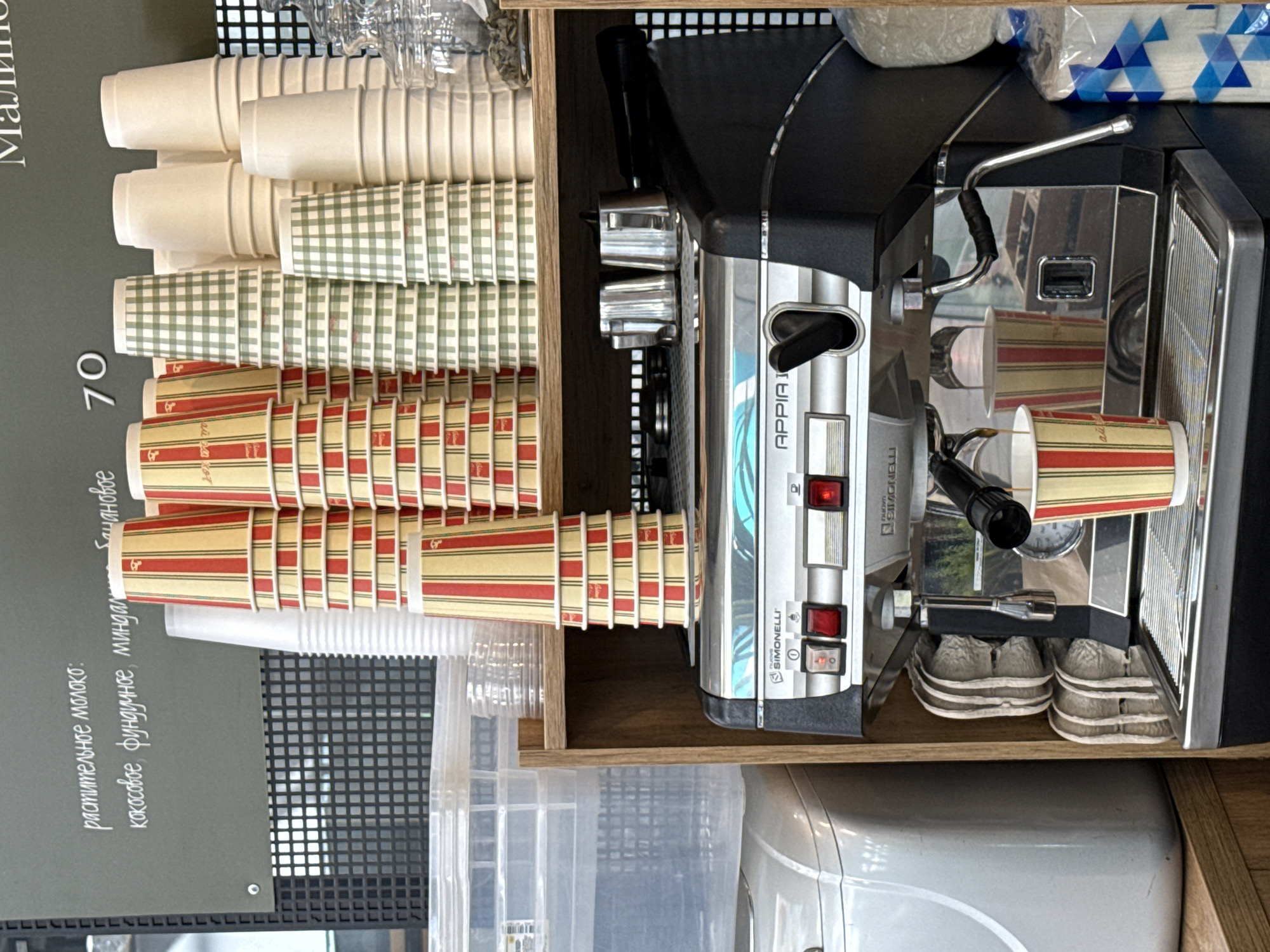 Here is a detailed description of the image:
**General Description:**
The image shows a close-up of a coffee preparation station, likely in a cafe or similar establishment. The scene is dominated by an espresso machine, stacks of disposable cups, and other equipment related to coffee making.
**Objects and Details:**
* **Espresso Machine:** A Simonelli Appia I espresso machine is prominently featured. It is silver-colored, with various levers, buttons, and a group head where coffee is extracted.
* **Disposable Cups:** Stacks of paper cups are visible. There are several different designs including plain white, green checkered and red and yellow striped
* **Countertop:** The station sits on a wooden countertop, with the shelf and additional supplies.
* **Other Equipment:** There are also containers likely holding various liquids or supplies.
**Environment:**
The scene is focused on the preparation area. The lighting appears to be artificial, typical of an indoor commercial setting.
**People and Actions:**
There are no people visible in the photograph. The scene appears to be prepared for service, indicating the location is a functional part of a business.
**Location:**
The location is most likely in a coffee shop or a cafe, a part of which would be in a city of Izhevsk, Russia.
