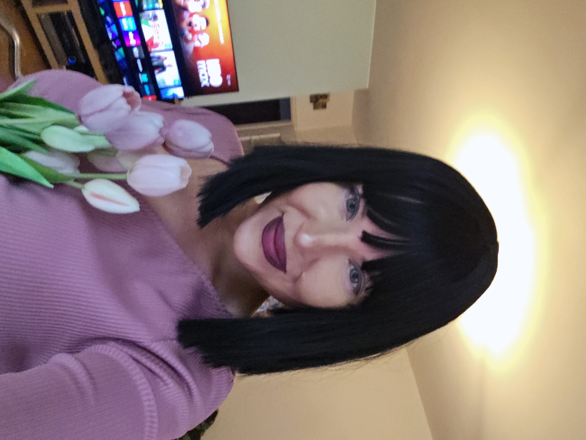 An indoor, sideways-oriented medium shot captures a person holding a bouquet of light pink tulips. The person, positioned in the center-left of the frame, has shoulder-length dark hair styled in a bob with bangs. They are smiling towards the camera, wearing dark pink or purple lipstick and visible eye makeup. Their top is a light purple, ribbed fabric.
The person is holding several light pink tulips with green stems and leaves, which are prominent in the foreground on the left side of the image.
In the background, a television screen is visible on the left, displaying a colorful streaming service interface with various show or movie thumbnails. The red text "MAX" is clearly discernible on the screen, suggesting it's the HBO Max streaming service. The rest of the background consists of light-colored walls, with a bright light source creating a warm glow on the wall to the right. The overall setting appears to be a home environment, likely a living room. The person's expression conveys happiness or contentment.