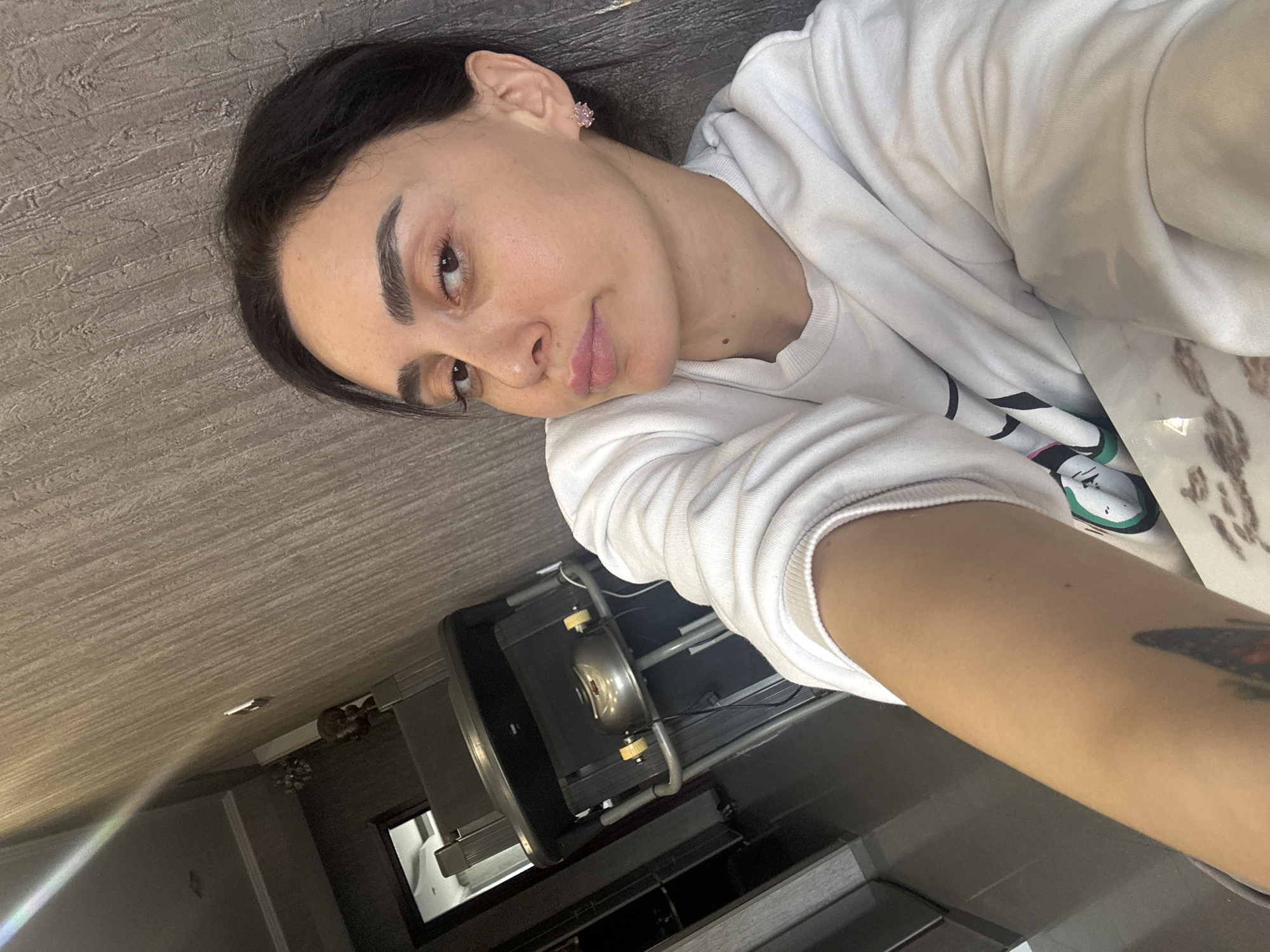 The image is a selfie of a young woman, likely taken indoors in a home setting in Tbilisi, Georgia. The woman is the central subject, with dark hair, dark eyebrows, and dark eyes. She gazes calmly towards the camera with a relaxed, slightly pensive expression, her head tilted and resting. She wears a plain white, long-sleeved sweatshirt. A small, light pink stud earring is visible in her right ear, and a subtle mole is present below her left eye. Her lips are naturally colored and slightly parted. A colorful tattoo, possibly a butterfly, is visible on her right forearm.
In the background, an indoor environment is depicted with a textured, light grey or brownish wall/floor surface. To the right of the woman, a metallic object, possibly a pot or a small appliance with two prominent yellowish-white knobs, sits on a dark-framed trolley or stand. Below this, dark furniture elements, potentially shelves or cabinets, are visible. A bright light source, possibly from a window or overhead fixture, creates a glare in the upper-left portion of the frame. The overall atmosphere is intimate and quiet. No discernible text is visible in the image. The time of day is not explicitly indicated, but the lighting suggests an interior setting with diffused illumination.