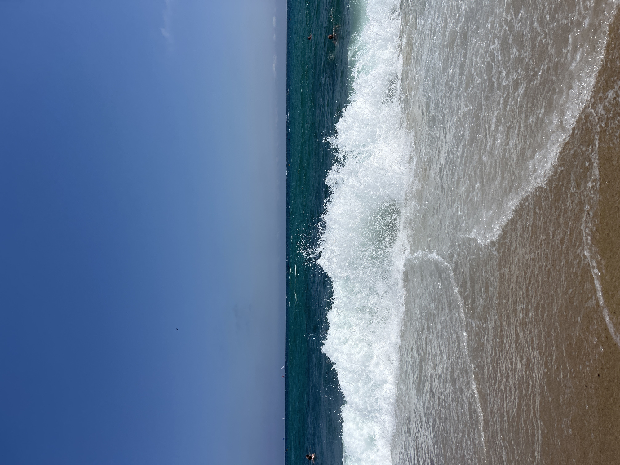 The image captures a dynamic ocean scene with a breaking wave in the foreground, set against a clear blue sky. The wave is prominent, with white foam and spray erupting as it crests. Gentle ripples of water are seen receding on the wet sand in the lower part of the frame. In the middle ground, the turquoise ocean stretches towards the horizon, dotted with several small sailboats and a few individuals swimming or wading in the water. A very faint smudge, possibly a distant cloud or a bird, is visible in the upper portion of the sky. The overall impression is of a sunny day at a beach. The presence of sailboats in the distance suggests a location where water sports are common, such as the Barceloneta Beach in Barcelona, Spain, known for its vibrant atmosphere and accessibility for various water activities.