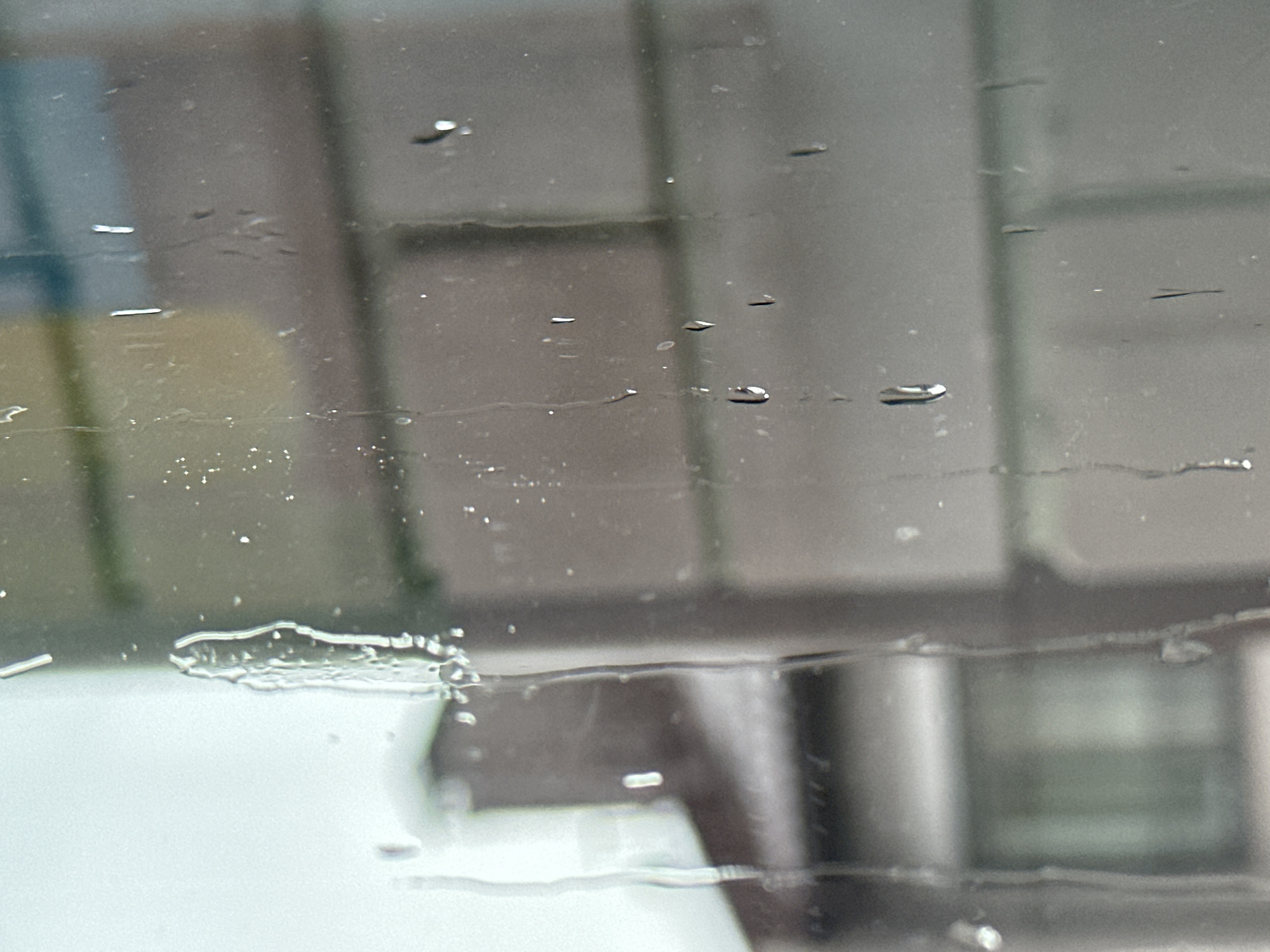 The image shows a close-up view of a window pane with water droplets and streaks. The background is blurred, but appears to be an urban environment with buildings and possibly trees. There are no people or discernible objects in the foreground. The lighting suggests it is daytime, possibly overcast or after rain. The location in Vladimir, Russia, is not evident from the visual information in the image.