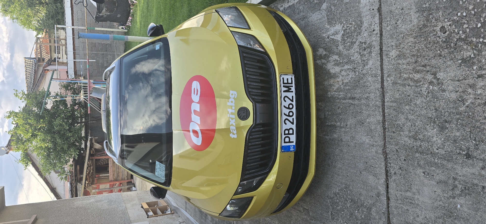 An overhead shot captures a yellow Skoda Octavia taxi parked on a concrete surface in Govedare, Bulgaria, during bright daylight. The car is the primary subject, displaying a large red oval logo with the white text "one" on its hood, accompanied by the smaller white text "taxi1.bg". The front license plate, visible at the bottom of the car, reads "PB 2662 ME" and features the "BG" country identifier in its blue EU strip.
The background shows elements of a residential or rural setting. To the left, a traditional-looking building with a tiled roof is partially visible, surrounded by lush green trees and foliage. Metal and wooden structures, possibly part of a clothesline or an outdoor utility area, are also present. The ground beneath and around the car is a textured concrete surface. The sky is partly cloudy, with reflections of clouds visible on the car's windshield. No people are visible, and the car appears to be stationary. The overall scene suggests a calm, daytime environment.