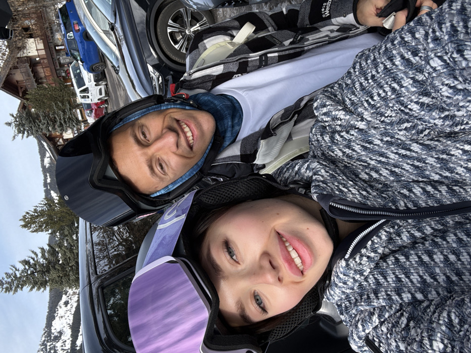 Two individuals, one female and one male, are depicted in a close-up selfie, smiling and looking directly at the camera. The female, positioned in the foreground on the left, wears a black ski helmet with purple-mirrored goggles pushed onto the helmet's brow. She is dressed in a blue and white patterned winter jacket. The male, positioned slightly behind her on the right, wears a black ski helmet over a blue neck gaiter or balaclava, a black and white plaid jacket over a white t-shirt, and what appears to be a light-colored backpack strap over his right shoulder. Both individuals exhibit expressions of contentment.
The background reveals an outdoor, snowy environment, indicative of a ski resort in Les Allues, France. Parked vehicles, including dark-colored SUVs, a blue compact car, and a white van, are visible behind the individuals. Further back, chalet-style buildings with wooden facades and sloped roofs are evident, interspersed with evergreen trees. Snow-covered terrain and distant mountains complete the scene under what appears to be daylight conditions.