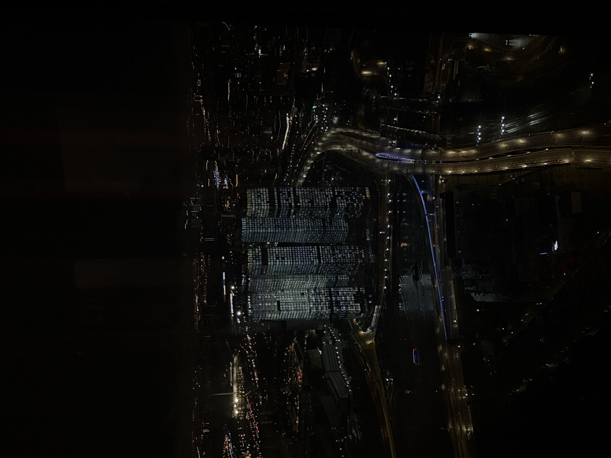 The image captures a nocturnal aerial view of a modern city skyline, with a particular focus on a cluster of illuminated skyscrapers. The buildings are rectangular and appear to be of varying heights, with many windows emitting a cool, white light, suggesting internal illumination. A body of water, likely a river or canal, is visible in the foreground, with reflections of the city lights shimmering on its surface. A boat, outlined by its lights, navigates the water.
A multi-lane highway, distinguished by its bright streetlights and the streaks of light from moving vehicles, curves through the scene, connecting different parts of the cityscape. Bridges, also illuminated, span across the waterway. In the background, a vast expanse of the city is visible, characterized by a dense network of smaller lights representing residential and commercial areas, stretching towards the horizon. Some larger, distinct structures with prominent lighting are scattered throughout the background, hinting at significant landmarks. The overall ambiance is one of urban activity continuing into the night, with the artificial lights creating a stark contrast against the dark sky. This scene is located in the city of Khimki, Russia, showcasing its developing infrastructure and urban landscape.