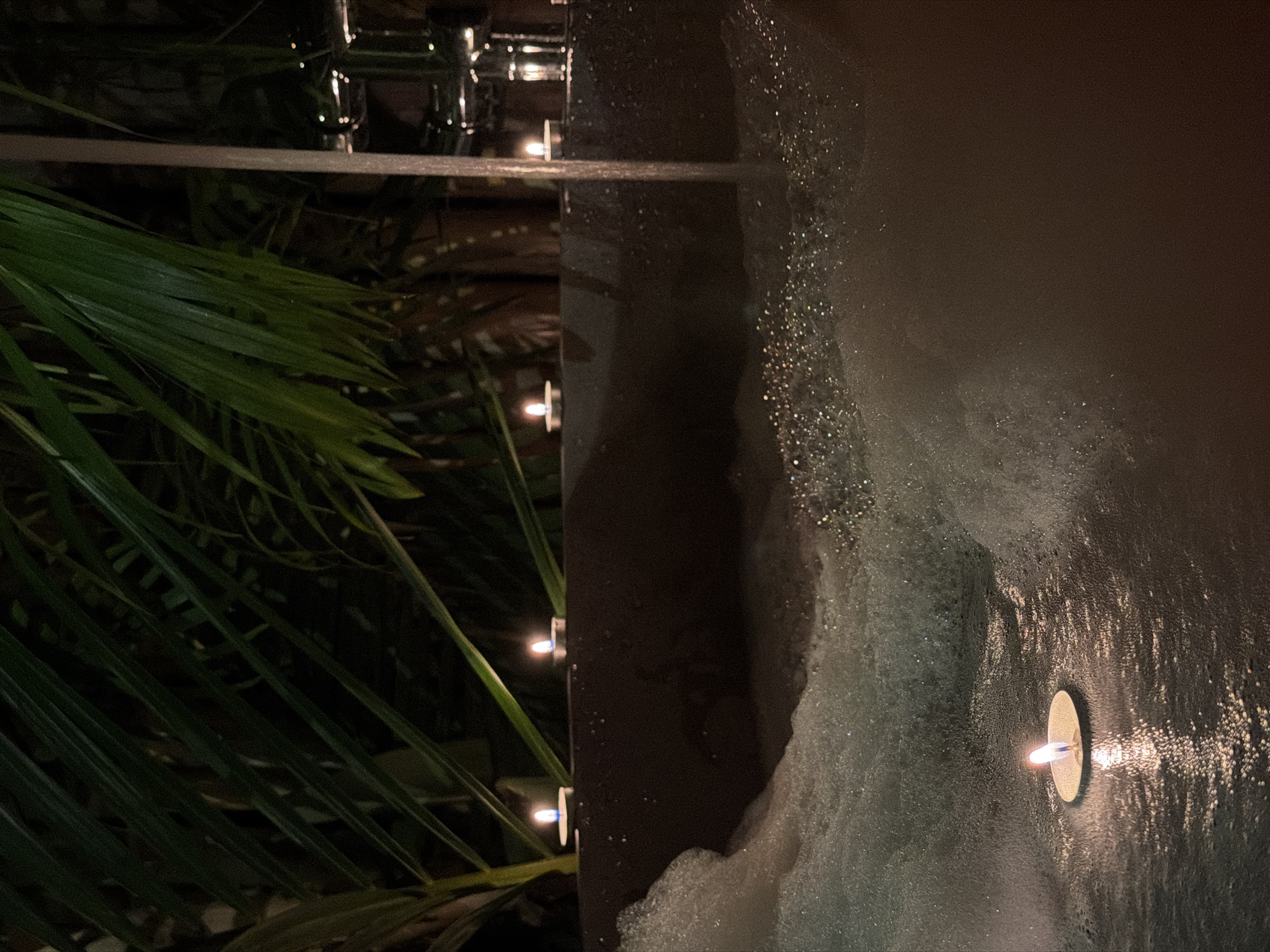 A lit tea candle floats in a bubble bath next to a person in a bathtub at night. The bathtub is dark brown and filled with white foamy bubbles. The surrounding environment is dark, with hints of palm leaves visible in the background, suggesting an outdoor or semi-outdoor setting. There are multiple other lit tea candles placed on the rim of the bathtub. The scene is illuminated by the soft glow of the candles and the ambient lighting. The location is identified as Baan Bo Phut, Thailand.