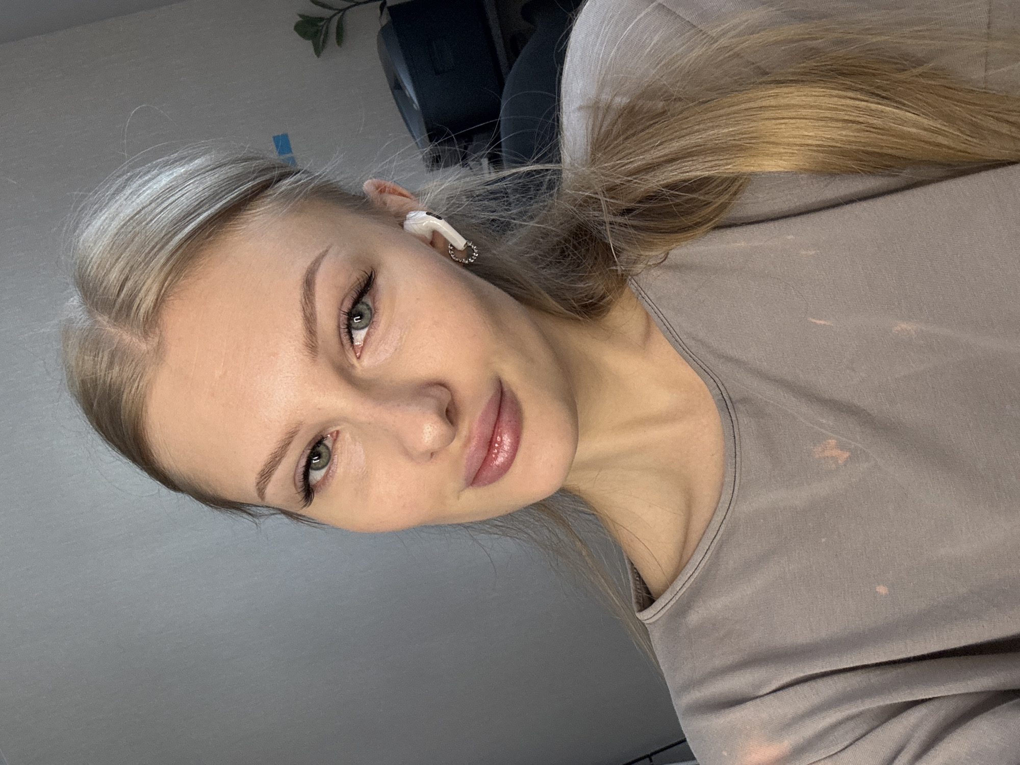 The image is a close-up, selfie-style portrait of a young woman with blonde hair and blue-green eyes. She is wearing a grey t-shirt and a white earbud in her right ear. She has on light makeup, including mascara and lip gloss. The background appears to be an indoor setting with a grey wall and some furniture. There is no visible indication of the city of Perm, Russia.