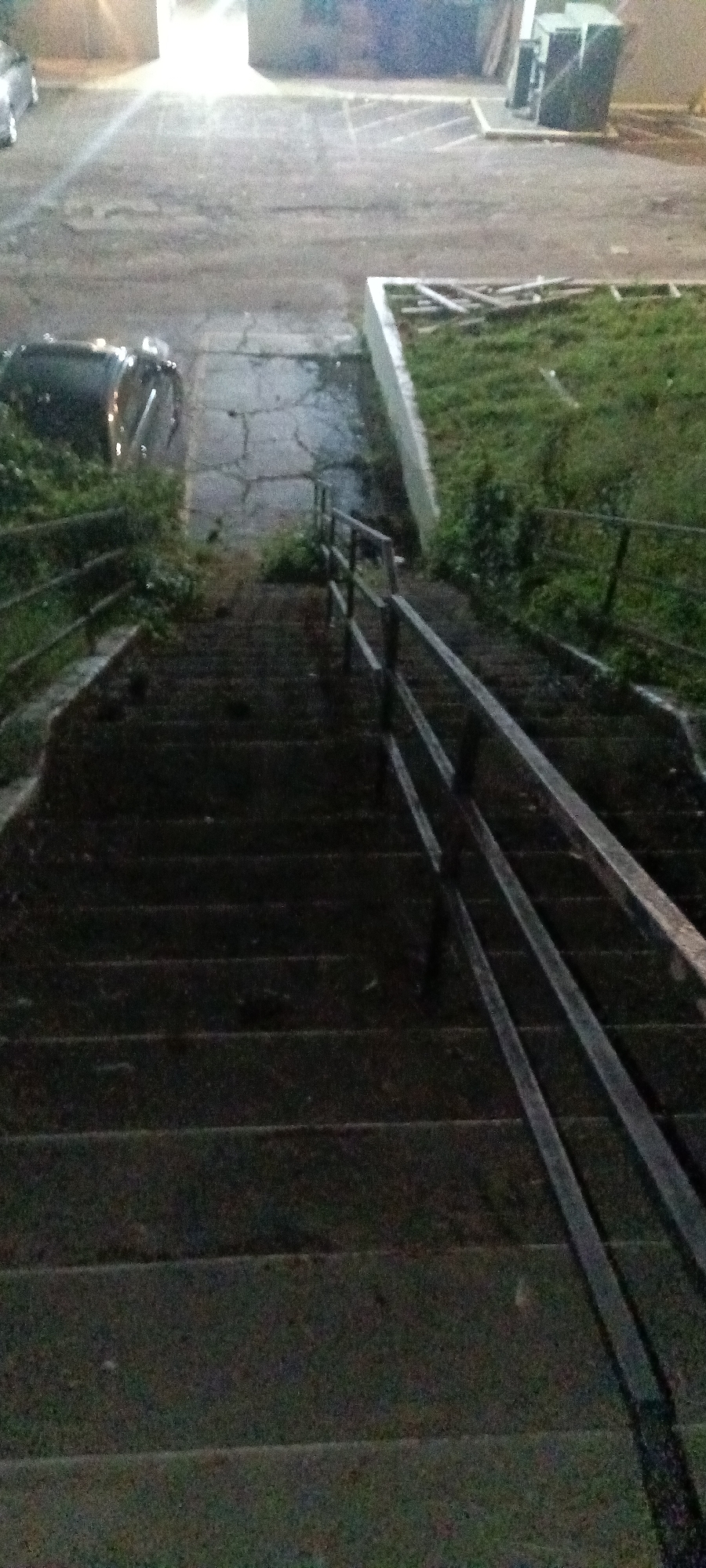 The image depicts a set of outdoor stairs leading down from a paved area to a lower level with grass and a railing. The stairs are made of stone or concrete and are dark in color, with visible wear. A metal railing runs along the right side of the stairs. Above the stairs, there is a cracked paved area with a car parked on the left. To the right of the stairs is a patch of grass bordered by a concrete retaining wall. In the background, there are buildings and a parking lot, illuminated by streetlights. The image was likely taken at night, given the dim lighting. The location is Fort Smith, United States of America.