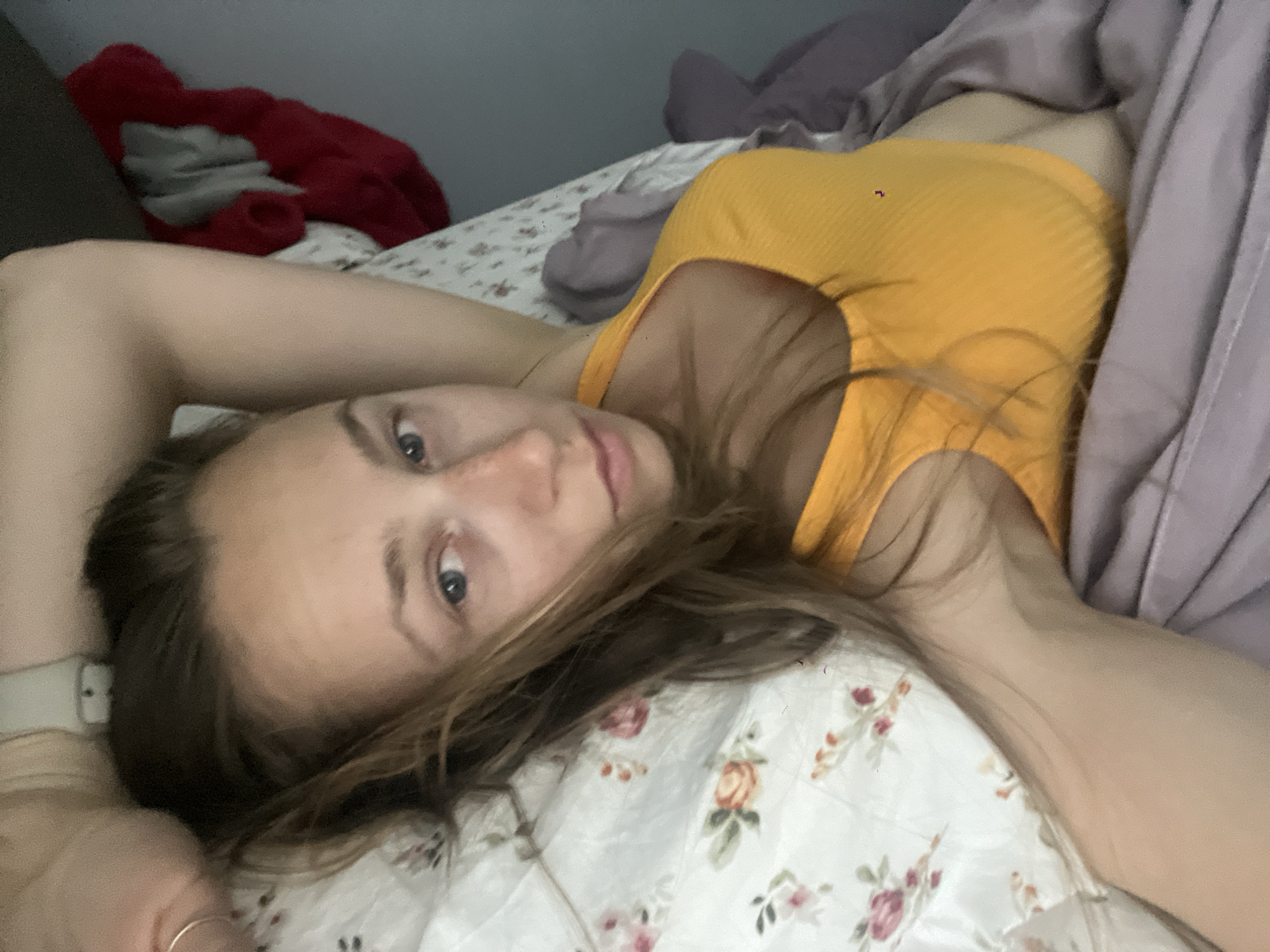 An individual is depicted lying on a bed in an indoor location within Bečići, Montenegro. The female subject is positioned on her back, with her head angled towards the left and her torso diagonally across the frame. Her gaze is directed towards the camera. She has light brown hair, partially resting on a white, floral-patterned comforter. She is wearing a ribbed, bright orange tank top. A light-colored wristwatch is present on her left wrist, and a simple silver-toned ring is visible on a finger of her right hand, which extends partially out of frame. A light purple sheet or blanket covers her lower body and is draped over the bed's right side. In the upper left background, a plain gray wall is visible, with a crumpled red fabric object and an additional dark gray or purple fabric item situated on the bed.