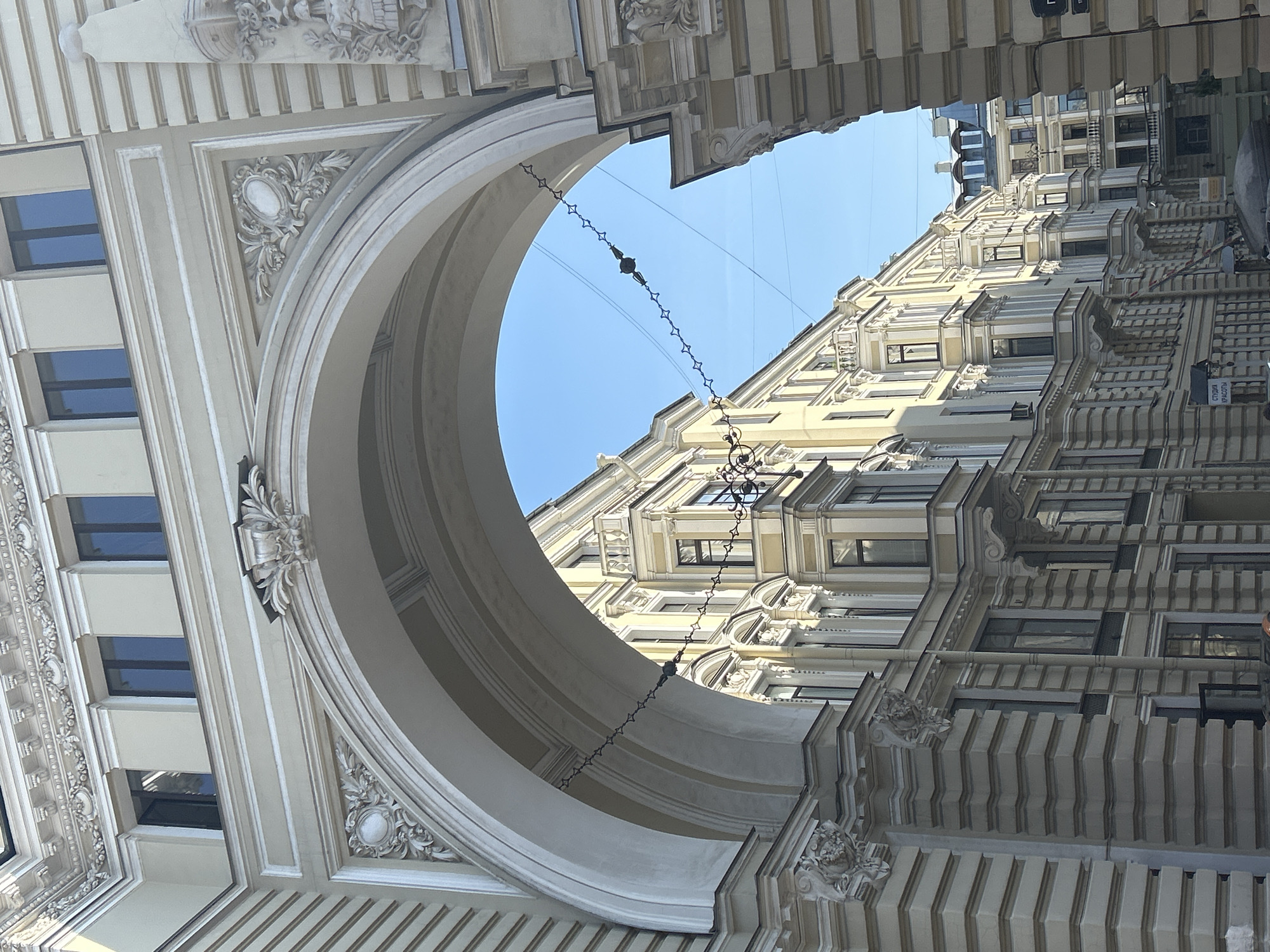 An ornate architectural archway frames a view of a street lined with historic buildings in an urban setting. The archway, constructed from light-colored stone or stucco, features intricate decorative carvings and moldings. Above the arch, the upper floors of a building are visible with several windows.
Through the arch, a narrow street unfolds, bordered by multi-story buildings with a consistent architectural style. These buildings are painted in light yellow and cream colors and feature numerous windows, bay windows, and balconies with decorative railings. The facades are adorned with classical architectural elements such as pilasters and cornices.
A series of decorative chains with star-shaped elements hang across the street, suspended from the buildings and archway. The sky is clear and blue, indicating daytime. The overall impression is one of old European city architecture, with a sense of grandeur and historical preservation.