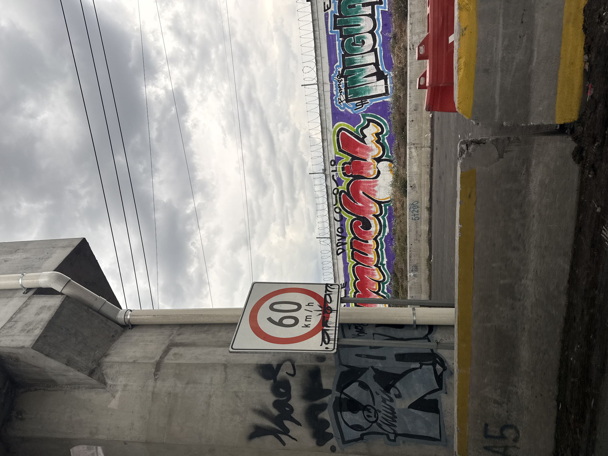 The image shows an outdoor urban scene in Valle de Chalco Solidaridad, Mexico. The foreground features a concrete structure, likely a bridge or overpass support, with a white PVC pipe attached. A speed limit sign indicating 60 km/h is affixed to the structure, marred by black graffiti. Additional graffiti covers the concrete surface below. In the background, a concrete barrier with colorful graffiti and barbed wire runs along the side of the road. A red and yellow traffic barrier sits on the right side of the barrier. Above, a cloudy sky is visible, with power lines stretching across the frame. The overall impression is of a run-down or neglected area.
