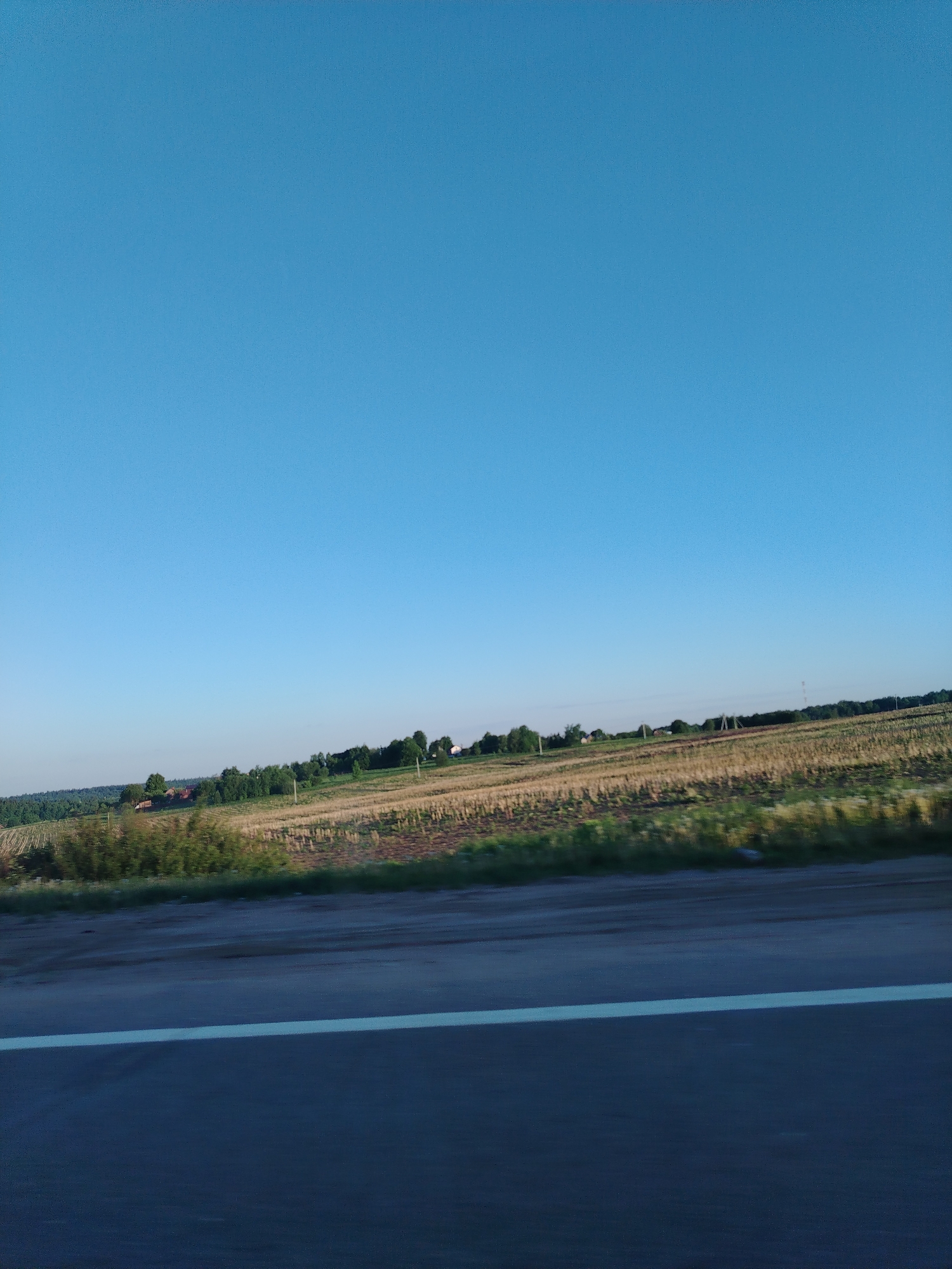 The image depicts a rural landscape. A clear blue sky dominates the upper portion of the frame. Below the sky, a field of harvested crops stretches towards a line of trees and foliage along the horizon. A road with a white lane marking cuts across the lower portion of the image, indicating movement. The location is unclear, there is no evidence to suggest that this is Mitino, Russia. The image lacks people.