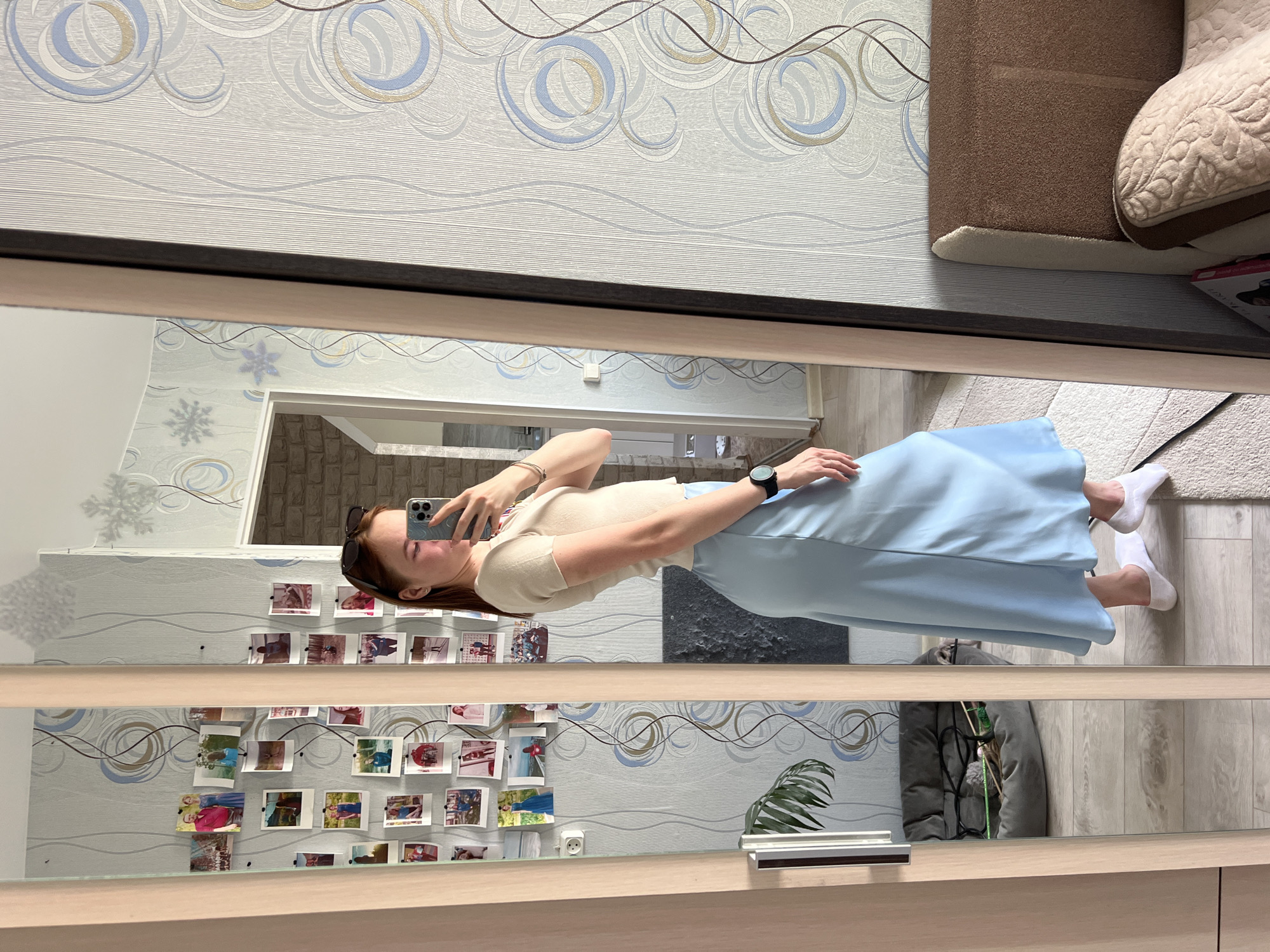A woman is standing in front of a mirror in a room. She is wearing a light beige short-sleeved shirt and a light blue skirt. She is holding a smartphone in her left hand, presumably taking a selfie. She is wearing a watch on her right wrist and sunglasses are perched on her head. In the mirror, on the wall behind her, is a decorative wallpaper featuring blue and gold circles. Small photographs are pinned to the wallpaper. An interior room with brick walls is visible through a doorway. A plant and a grey coat are visible in the lower portion of the mirror. A sofa, a rug, and wood flooring are in the room where the woman is standing. An electrical outlet is visible in the mirror. The location is Chaikovsky, Russia.