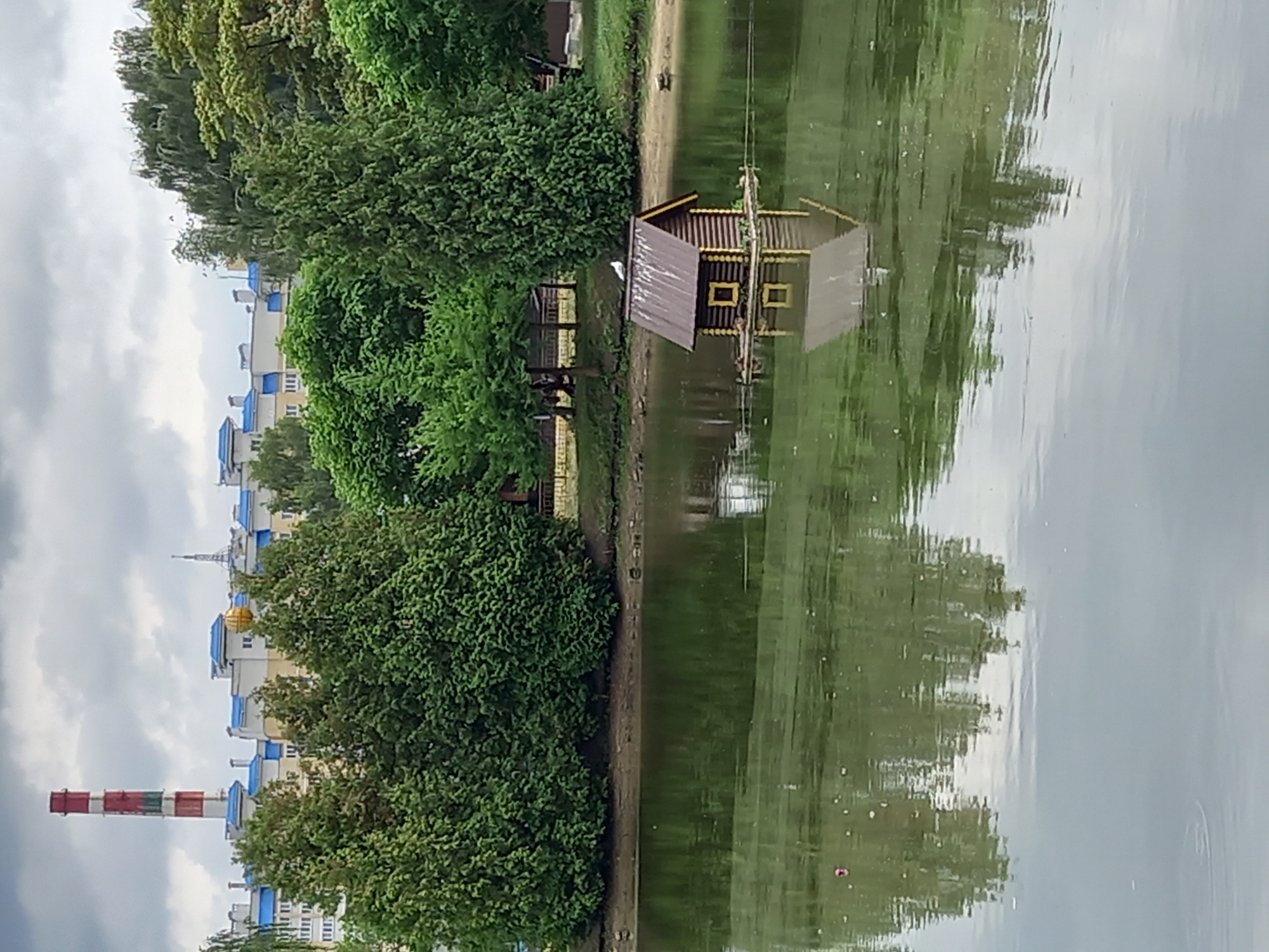 This image depicts a serene waterscape with a small, wooden house seemingly floating on the surface of a calm body of water. The house has a dark, ribbed exterior and a sloped, darker roof, with two yellow-framed windows visible. Its reflection is clearly mirrored in the water below, creating a symmetrical effect.
Surrounding the water is a lush, green landscape with dense trees and bushes on the left bank. Further in the background, on the left side of the image, a row of buildings with blue roofs and white facades can be seen, along with a tall, striped industrial chimney in shades of red and white. This suggests an urban or semi-urban environment adjacent to a natural water feature.
The sky appears overcast with soft, diffused light, indicating it might be a cloudy day or close to dawn or dusk. The overall mood is tranquil and peaceful, with the stillness of the water and the greenery contributing to a sense of calm. There are no visible people or specific activities taking place, making it a still-life composition focusing on the interplay of architecture, nature, and reflection. The location context provided, Brest, Belarus, helps to ground the scene within a specific geographical area.
