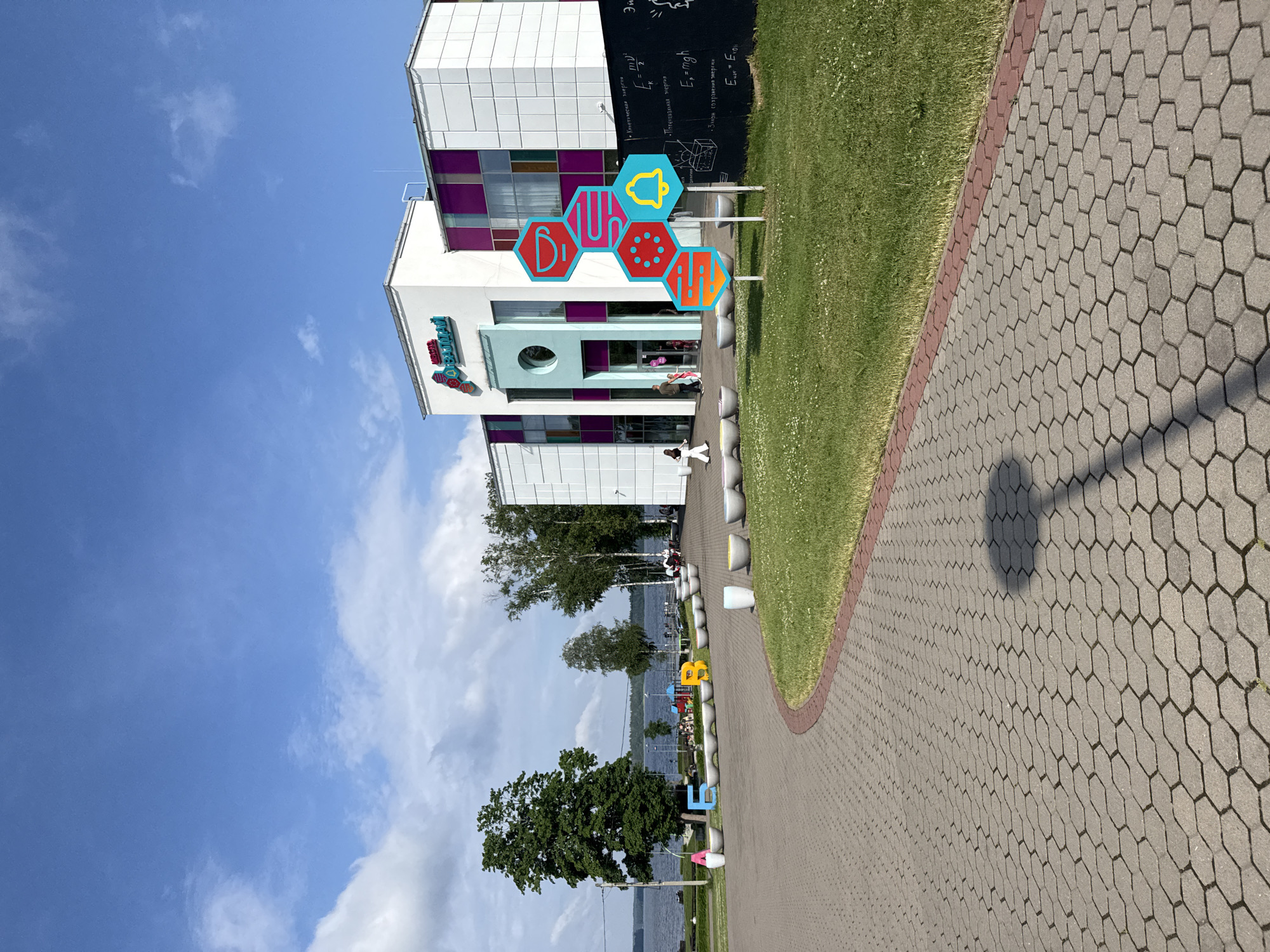 In Valday, Russia, a modern building with a white facade and purple accents stands under a blue sky with scattered clouds. In front of the building, a large, colorful honeycomb-like sculpture with icons of a bell, a coffee cup, and other symbols is displayed on the grass. A blackboard with mathematical equations is visible to the right of the sculpture. To the left of the building, a paved walkway leads towards the foreground, bordered by a grassy area and several low concrete planters. Further back, large colorful letters spelling "БЕГ" (meaning "run" in Russian) are visible on the grass, with a lake and trees in the background. Several people are walking and sitting in the area.
