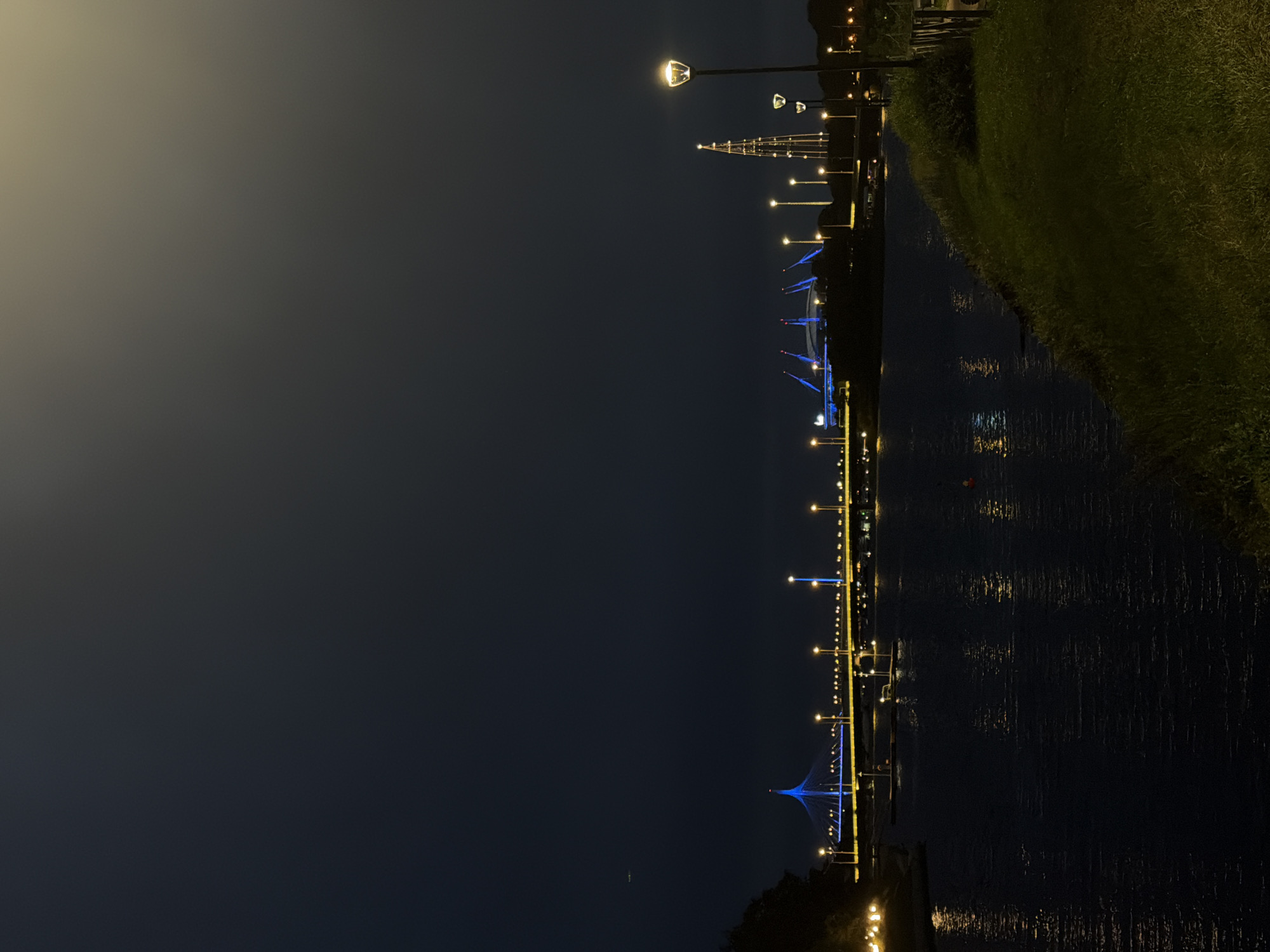 This is a nighttime, landscape orientation photograph taken outdoors. The scene depicts a waterfront area illuminated by artificial lights. A dark body of water occupies the right side of the frame, reflecting the lights from the opposite shore. On the left side of the water, a walkway or embankment is lined with lampposts that cast a warm, yellow glow. Further along this embankment, a suspension bridge is visible, distinguished by its vibrant blue illumination. Beyond the bridge, a dark treeline or land mass is suggested. The sky is a deep, dark blue, characteristic of nighttime. The overall mood is serene and somewhat atmospheric due to the contrasting lights and darkness. There are no visible people or text in the image.