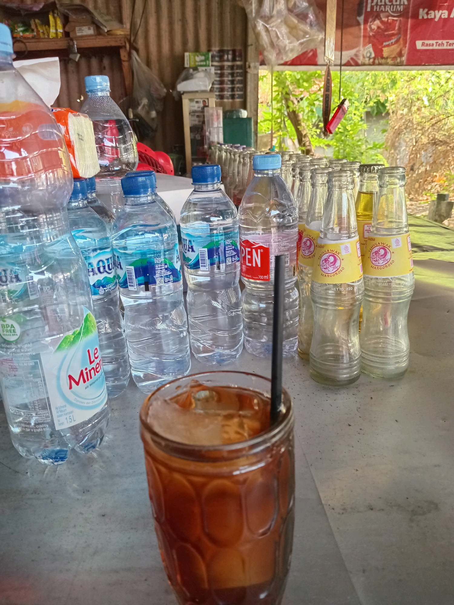 In Kembangan, Indonesia, a glass of iced tea sits on a table in front of a row of plastic water bottles and glass bottles containing a yellow liquid. The table is covered with a light-colored tarp. In the background, a corrugated metal wall is visible, along with shelves stocked with various goods, including bags of snacks and what appear to be cigarette packs. The scene suggests an outdoor or semi-outdoor stall or shop, possibly a warung. The overall lighting indicates it is daytime, with natural light filtering in from the right side.