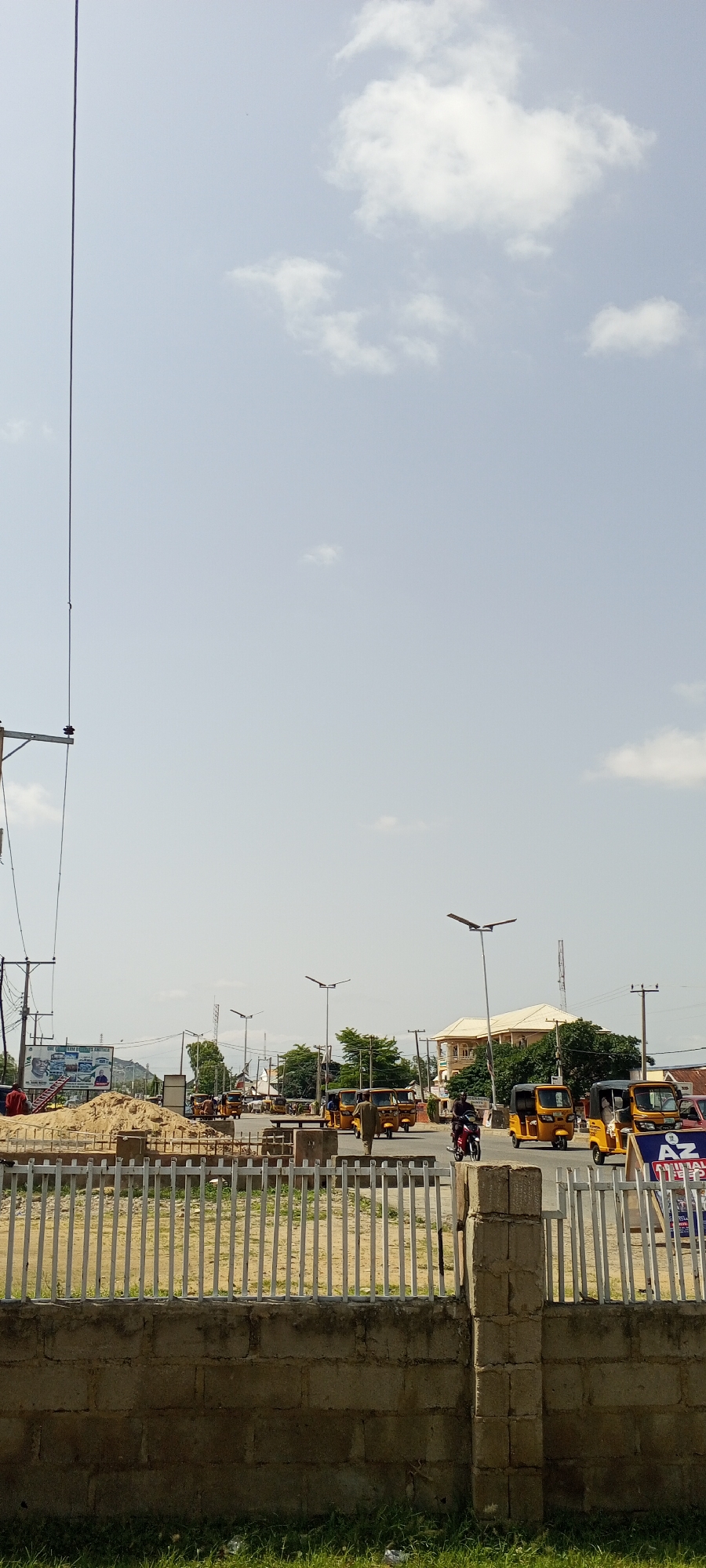 The image depicts a street scene in Jalingo, Nigeria. In the foreground, a low, weathered brick wall topped with a white metal fence stretches across the frame. Beyond the fence, a construction site is visible with a large mound of sand and some scaffolding. In the middle ground, a street is lined with numerous yellow, three-wheeled auto-rickshaws, commonly known as Keke Napep, some of which are stationary and others are in motion. A few people are visible, including a man walking away from the camera on the street, and another person riding a motorcycle. In the background, there are buildings with light-colored walls and roofs, trees, and utility poles with wires. The sky is clear with scattered white clouds. The overall impression is of a busy urban environment during daylight hours.