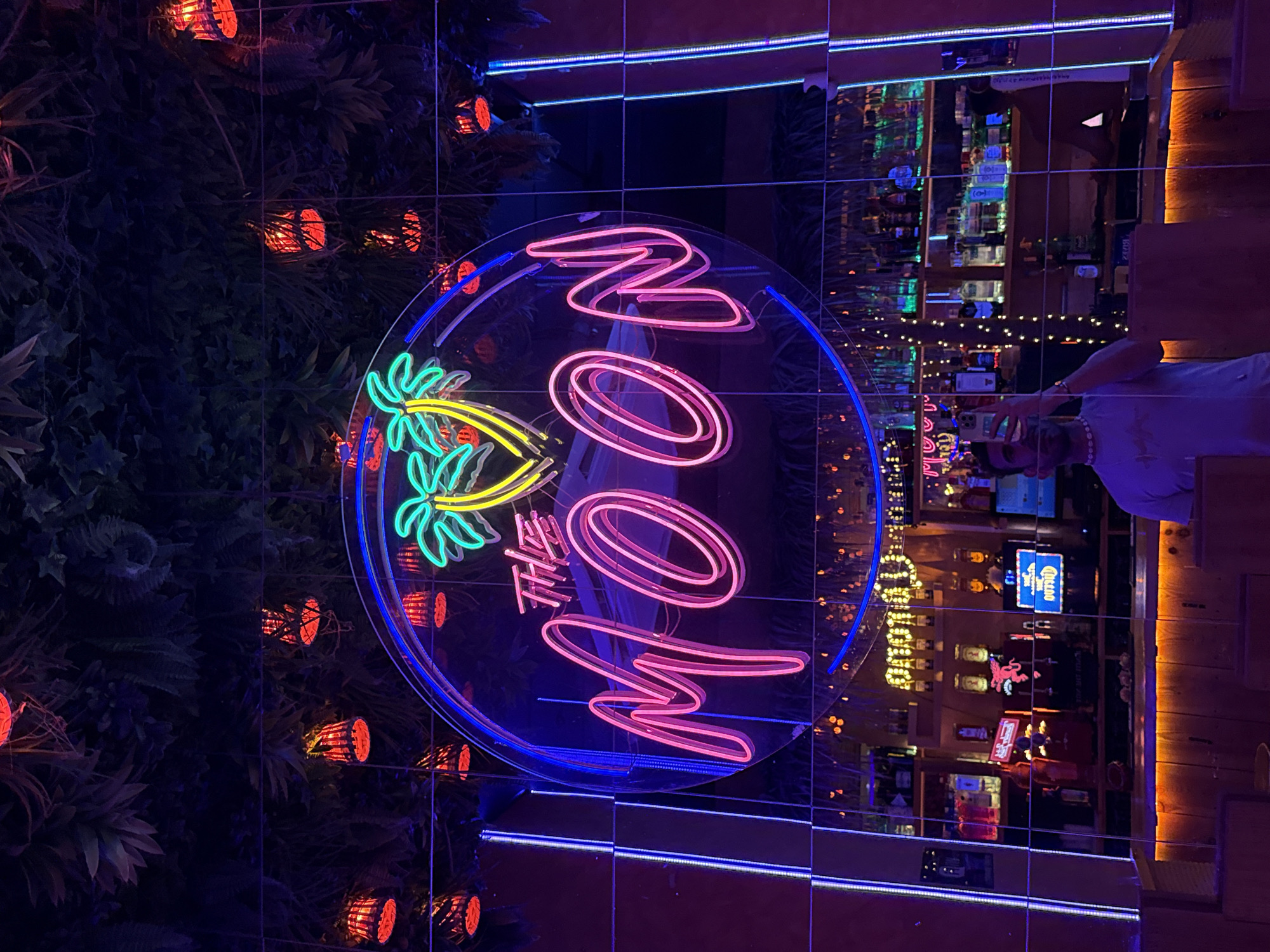 The image depicts a neon sign in a dimly lit establishment. The sign, in cursive pink neon, reads "THE MOON" and is surrounded by a blue neon circle. Above the sign, artificial palm leaves and red lanterns hang from the ceiling. The sign is reflected in a mirrored wall, behind which a bar with shelves of bottles is visible. A person is taking a selfie in the reflection. The establishment appears to be a bar or nightclub, possibly in Barcelona, Spain, given the context of the question.