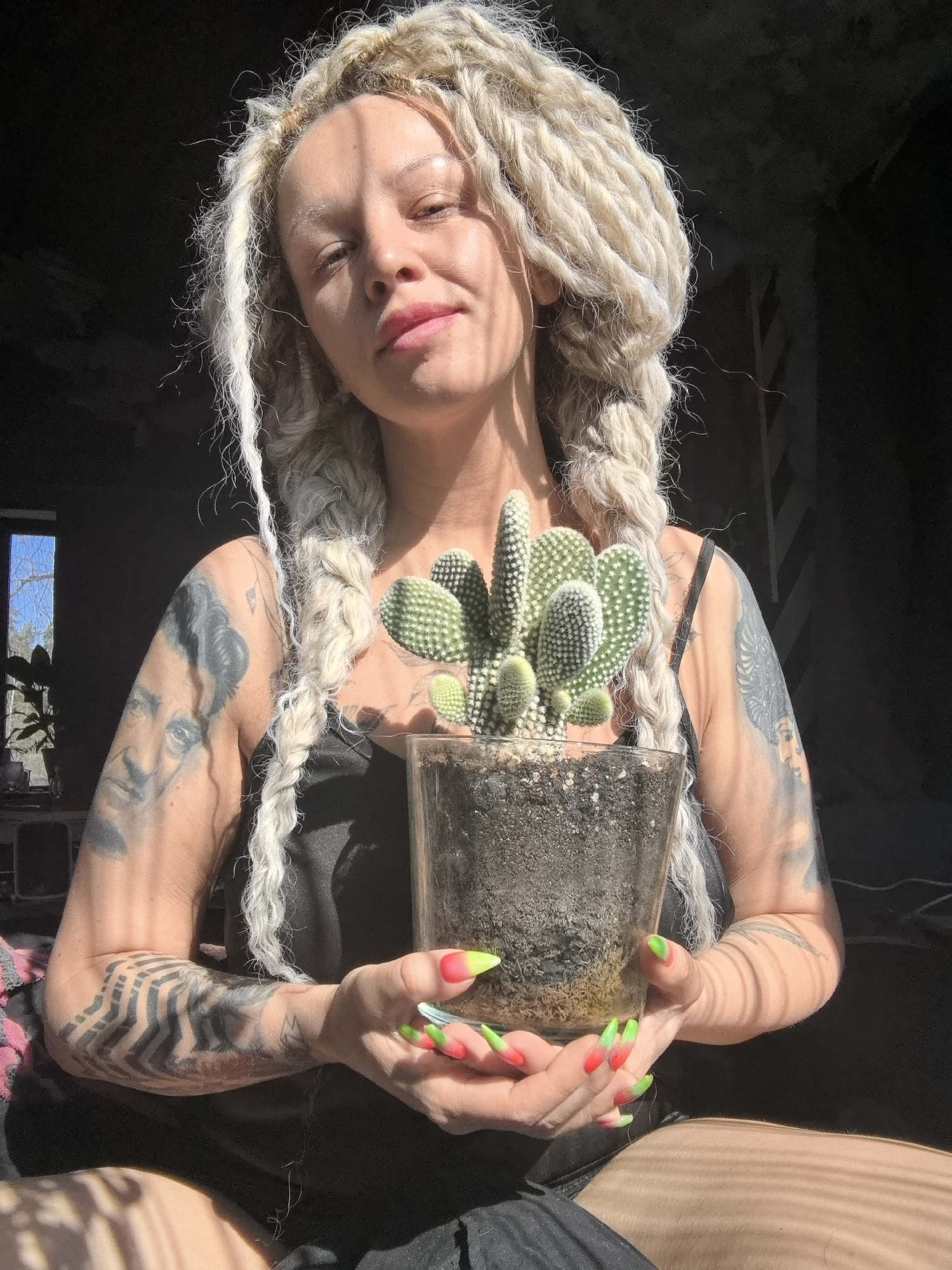 A woman with long, light-blonde dreadlocks and extensively tattooed arms is depicted indoors, holding a potted cactus. She wears a black tank top and looks directly at the viewer with a soft, gentle smile. Her nails are long and pointed, featuring a vibrant red-to-neon-green ombre design.
The woman's left upper arm displays a large tattoo that appears to be a portrait, while her right forearm is covered with a bold, geometric, maze-like pattern. She is holding a small, healthy *Opuntia microdasys* (bunny ear cactus) in a clear glass pot, revealing the dark soil within.
Strong, direct sunlight streams in from an unseen window, casting prominent striped shadows across her face, arms, and leg, indicating a clear, sunny daytime setting. The background is predominantly dark, appearing to be an interior room. To the left, a window provides a view of an outdoor scene featuring bare trees against a bright sky, suggesting the time of year might be winter or early spring in Noviy, Russia. The overall mood is calm and serene. There is no visible text in the image.