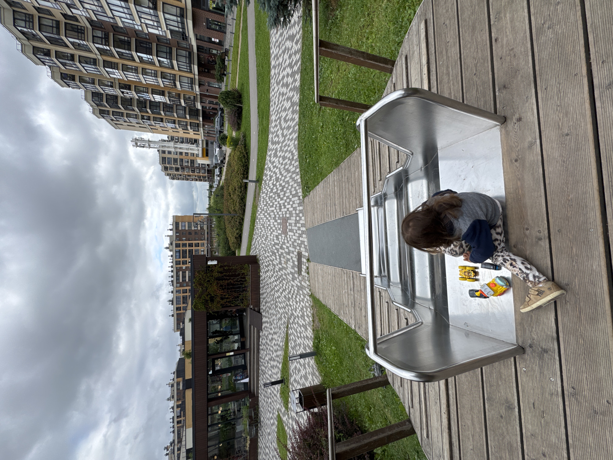 A child with long brown hair sits on a wooden deck at the top of a metal slide, facing away from the camera. They are wearing patterned leggings, a grey sweatshirt, and brown shoes. The child is playing with three toy vehicles on the slide's surface: a yellow truck, a yellow and red train engine, and a black object. The slide is part of a playground in a residential area. In the background, modern apartment buildings with many windows dominate the skyline under a cloudy sky. A building with large glass windows, possibly a cafe or store, is visible to the left. The ground is a mix of paved pathways and green grass. The scene appears to be in Novoselye, Russia.