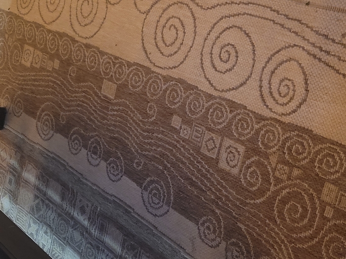 The scene presents an angled, close-up view of a woven textile, consistent with a floor rug or mat. The textile features multiple vertical bands of contrasting patterns and tones in a palette of browns, beiges, and creams. The leftmost visible band exhibits geometric square and X-shaped motifs. Adjacent to it, a wider band displays a complex arrangement of wavy lines, smaller square elements, and spirals. To the right, a prominent band showcases larger, distinct spiral patterns against a lighter background. The weaving appears to be a coarse, flat-weave texture. Uneven lighting is present, casting a slight shadow over the upper left portion of the textile. No individuals, additional objects, specific actions, or interactions are visible. The surrounding environment is not discernible beyond the textile itself. The image does not contain data to specify the location within Κωστήτσι, Greece.