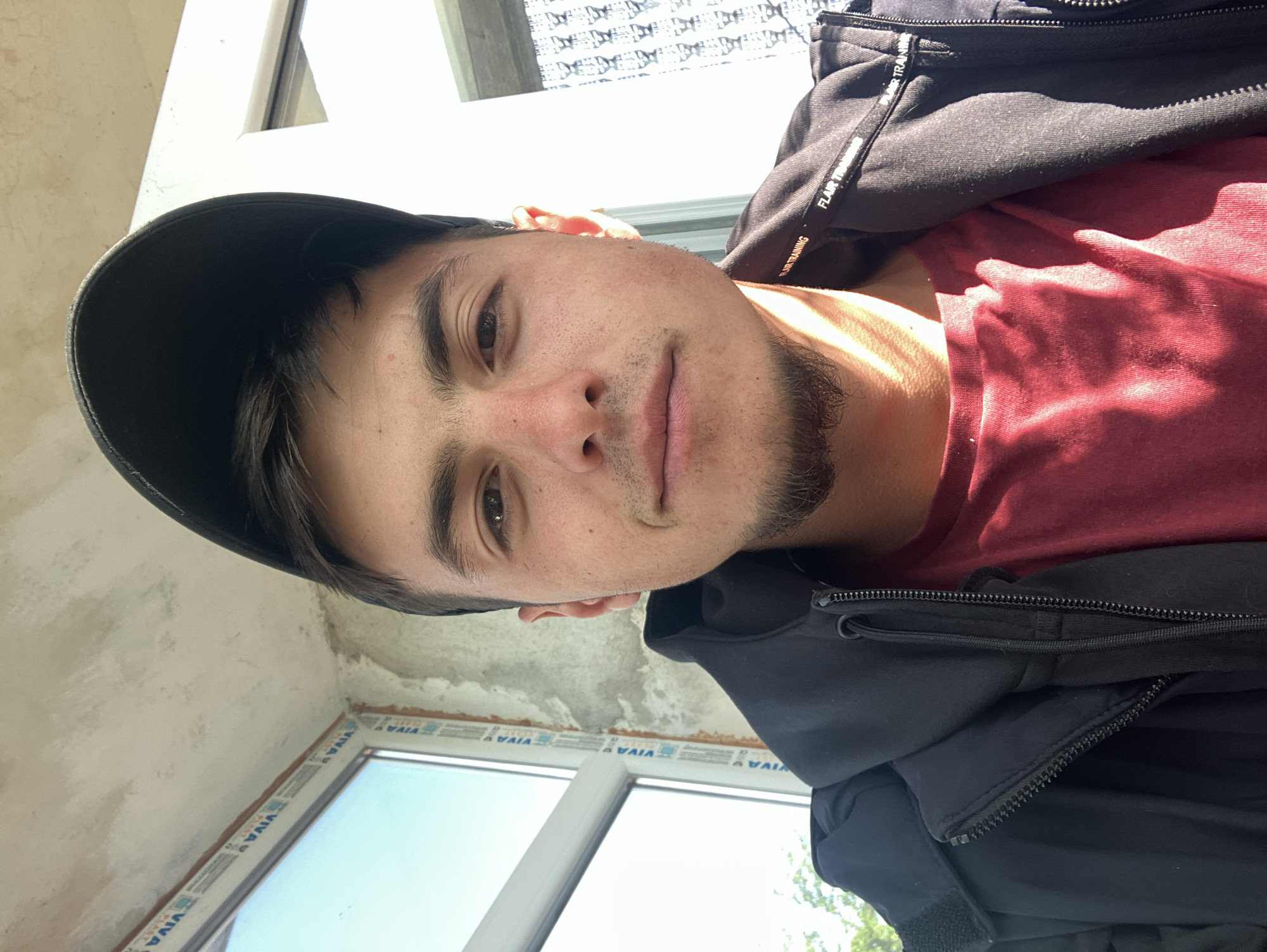 The image shows a man wearing a black baseball cap, a red t-shirt, and a black zip-up jacket. He appears to be indoors, near a window with a "VIVA PLAST" frame. The walls of the room appear to be under construction or renovation. While the image contains potential geographical indicators, a definite location in Krastevich, Bulgaria cannot be ascertained with certainty. The man is looking directly at the camera.